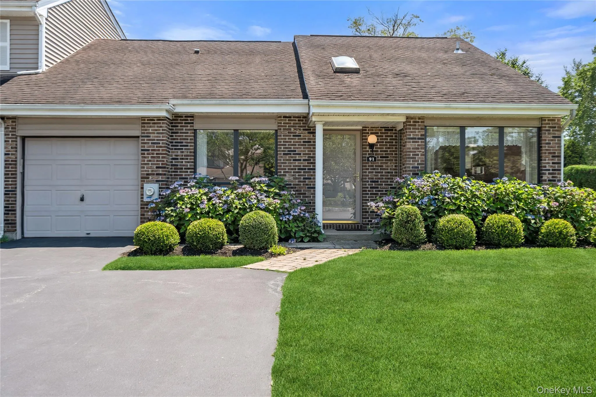 Emory Ranch style Brick end unit featuring a single-car garage, mature landscaping, a paved driveway, a skylight, and a walkway leading to the entry Emory Ranch style Brick end unit featuring a single-car garage, mature landscaping, a paved driveway, a skylight, and a walkway leading to the entry