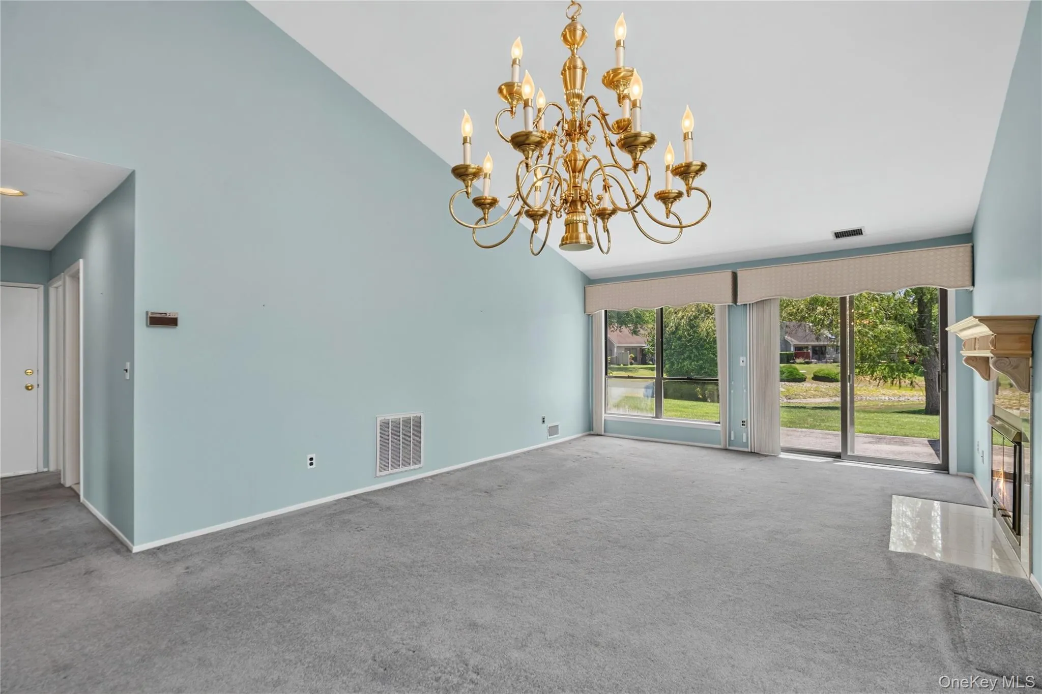 Vaulted ceilings and expansive windows frame the living area, featuring a brass chandelier and a fireplace with a light-toned hearth Vaulted ceilings and expansive windows frame the living area, featuring a brass chandelier and a fireplace with a light-toned hearth