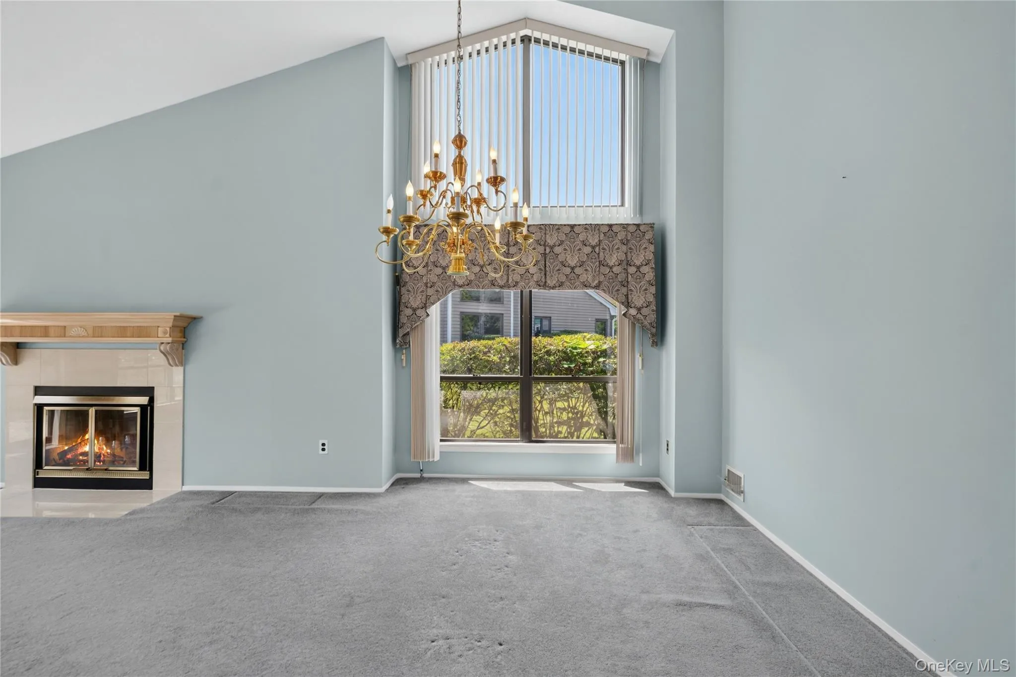 Vaulted ceilings featuring a chandelier, a fireplace with a stone-look mantel, and a large window with vertical blinds Vaulted ceilings featuring a chandelier, a fireplace with a stone-look mantel, and a large window with vertical blinds