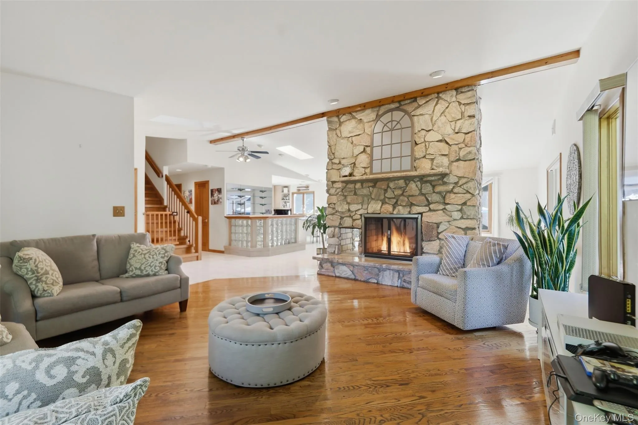 Spacious living area featuring stone fireplace with hearth, wood-finish flooring, exposed wood beam, and staircase with wood railing Spacious living area featuring stone fireplace with hearth, wood-finish flooring, exposed wood beam, and staircase with wood railing