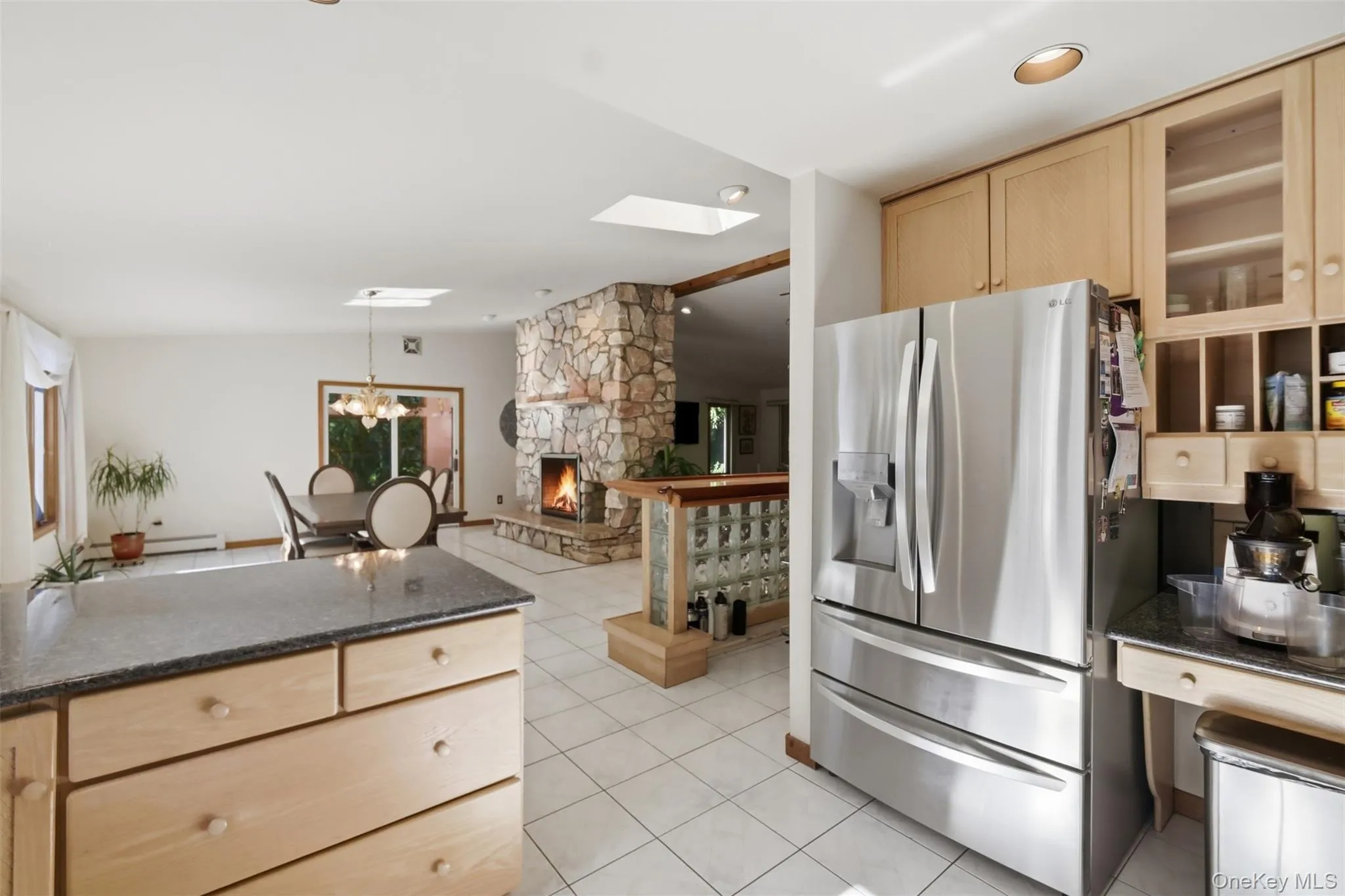 Spacious kitchen featuring a large island with a dark countertop, light wood cabinetry, and white tile flooring Spacious kitchen featuring a large island with a dark countertop, light wood cabinetry, and white tile flooring