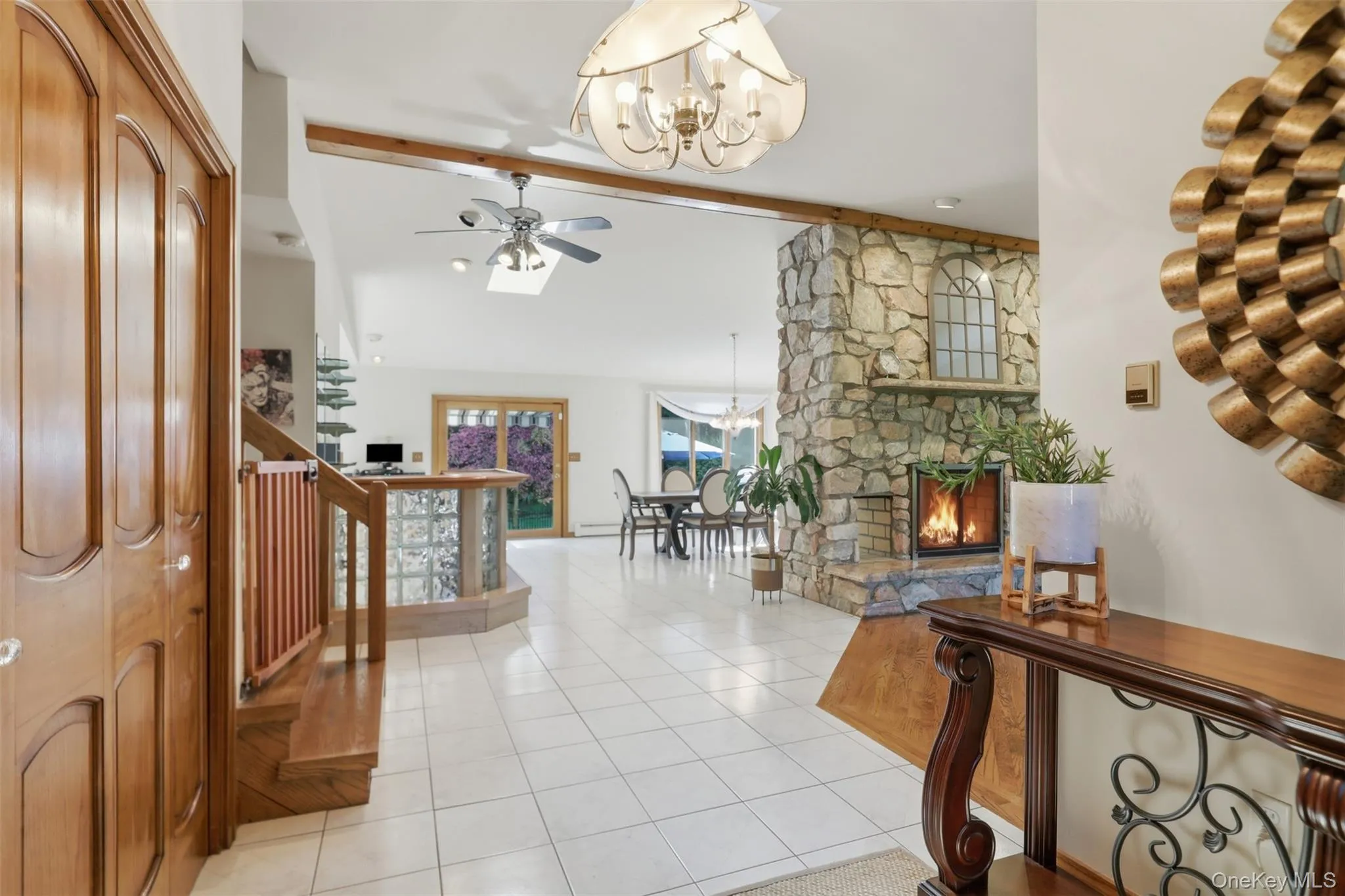 Stone-clad fireplace with hearth, vaulted ceiling with exposed beam, white tile flooring, natural wood staircase, and glass block partition Stone-clad fireplace with hearth, vaulted ceiling with exposed beam, white tile flooring, natural wood staircase, and glass block partition