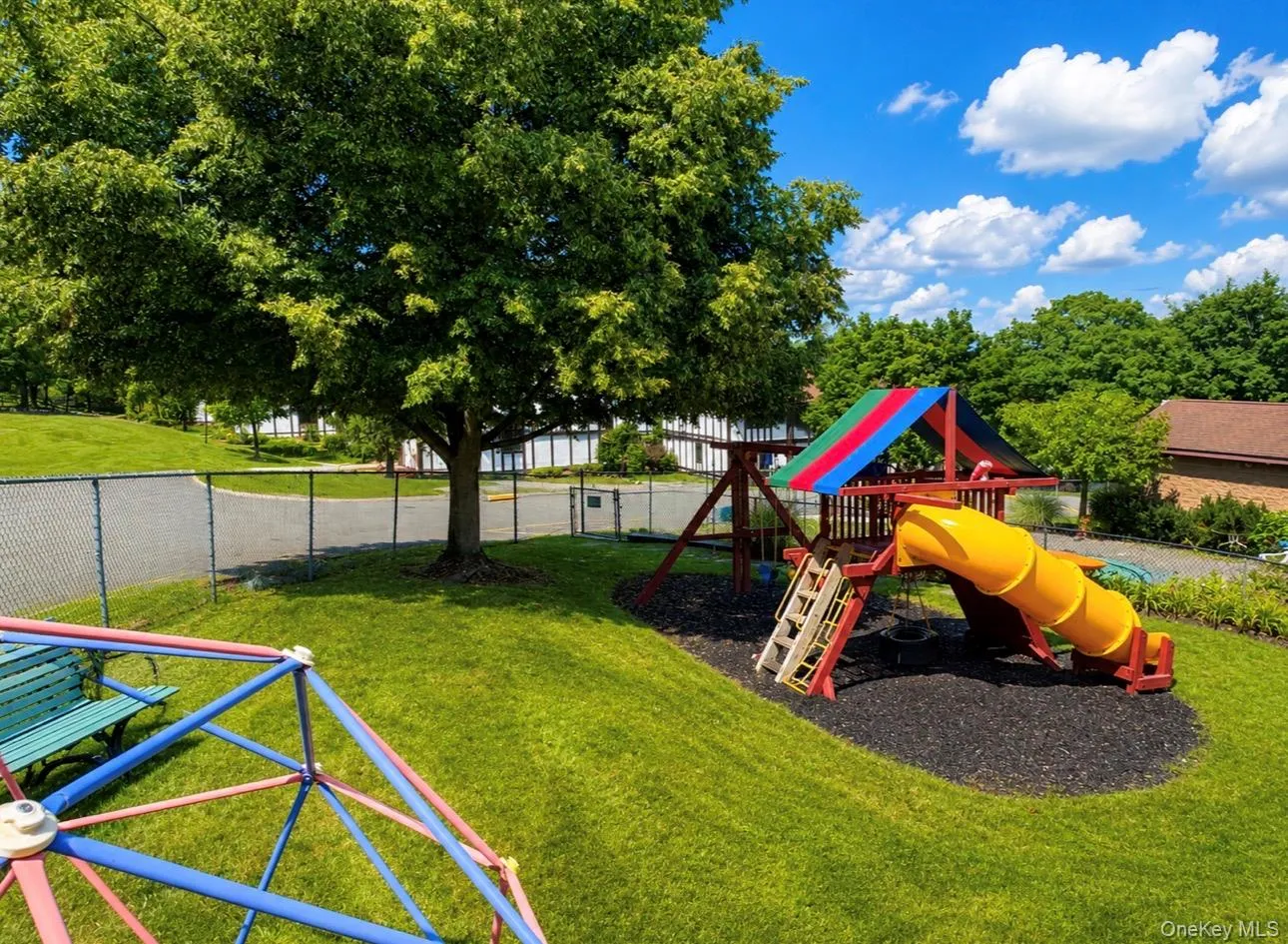 Expansive green space featuring a play structure with a tunnel slide, swings, and rock climbing wall, surrounded by mature trees and manicured lawn Expansive green space featuring a play structure with a tunnel slide, swings, and rock climbing wall, surrounded by mature trees and manicured lawn
