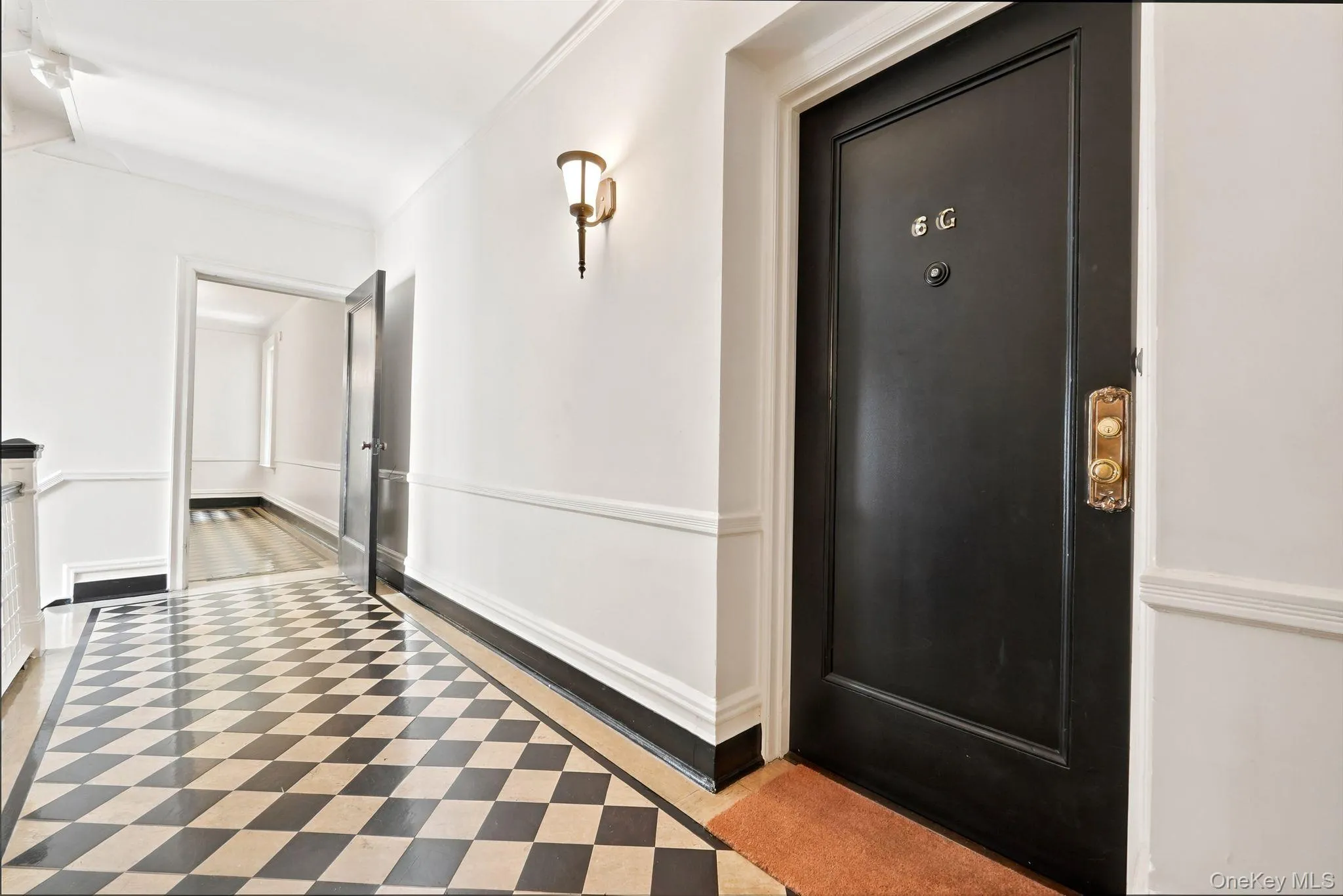 Entryway featuring checkered flooring, a black front door with brass hardware, a wall sconce, white walls with wainscoting, and a framed doorway Entryway featuring checkered flooring, a black front door with brass hardware, a wall sconce, white walls with wainscoting, and a framed doorway