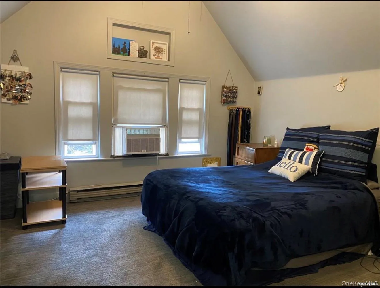 Vaulted ceiling bedroom featuring three windows, a window-mounted air conditioning unit, baseboard heating, and gray carpeting Vaulted ceiling bedroom featuring three windows, a window-mounted air conditioning unit, baseboard heating, and gray carpeting