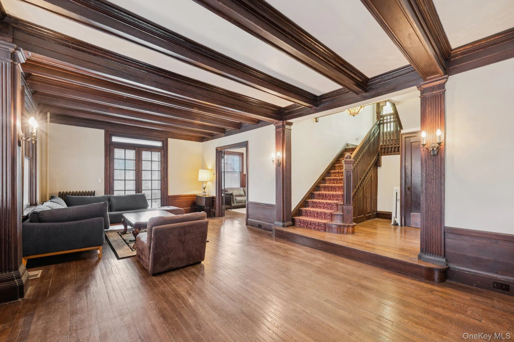 Living room featuring ornate columns, beamed ceiling, wainscoting, stairway, and wood-type flooring Living room featuring ornate columns, beamed ceiling, wainscoting, stairway, and wood-type flooring