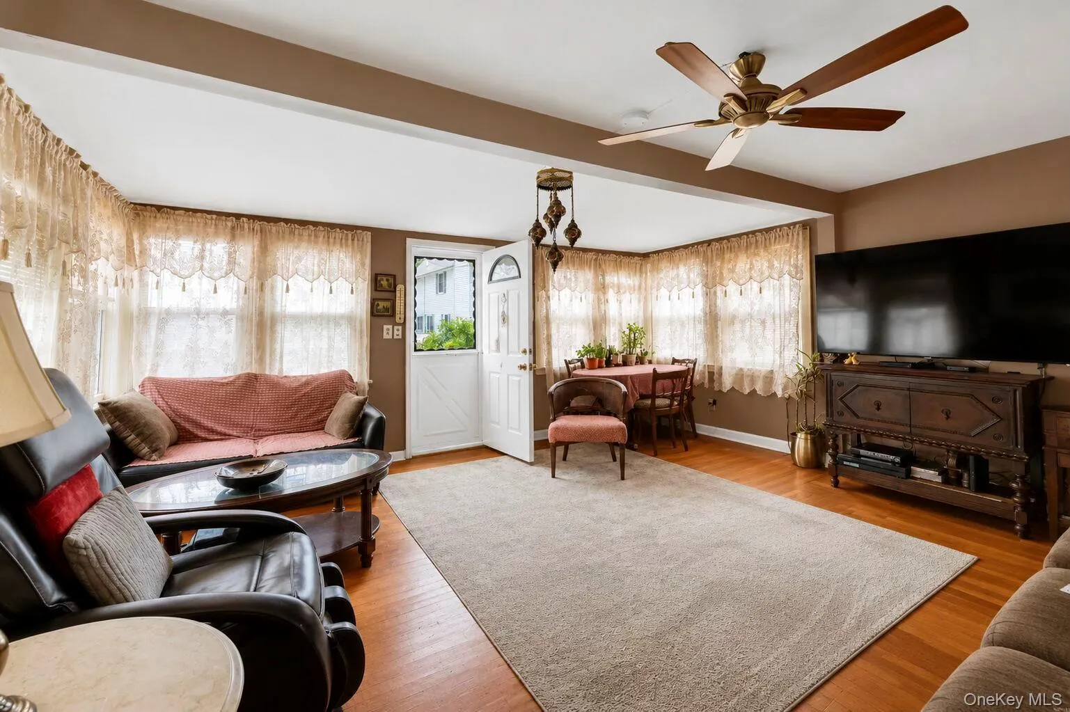 Spacious living area featuring wood-finish flooring, a ceiling fan with integrated lighting, and multiple windows Spacious living area featuring wood-finish flooring, a ceiling fan with integrated lighting, and multiple windows