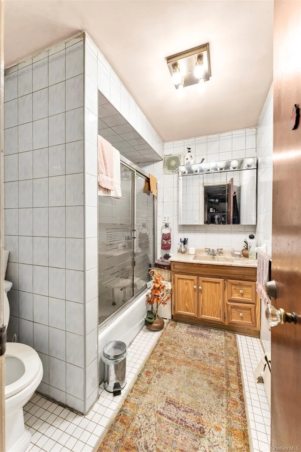 Bathroom featuring white square wall and floor tiling, a single-sink wood vanity with a light-toned countertop, and a shower-tub combination with sliding glass doors Bathroom featuring white square wall and floor tiling, a single-sink wood vanity with a light-toned countertop, and a shower-tub combination with sliding glass doors