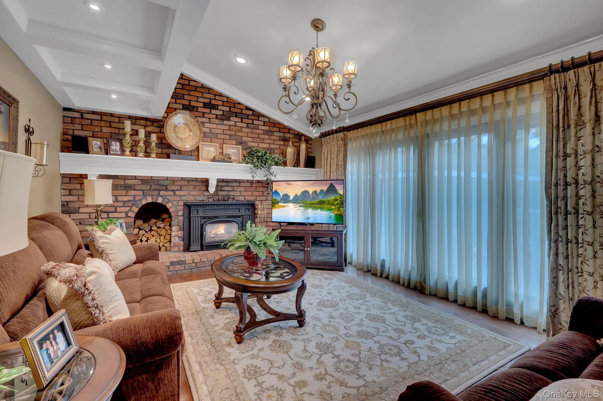 Living area featuring a brick accent wall with a fireplace and mantel, tray ceiling with recessed lighting, and a chandelier Living area featuring a brick accent wall with a fireplace and mantel, tray ceiling with recessed lighting, and a chandelier