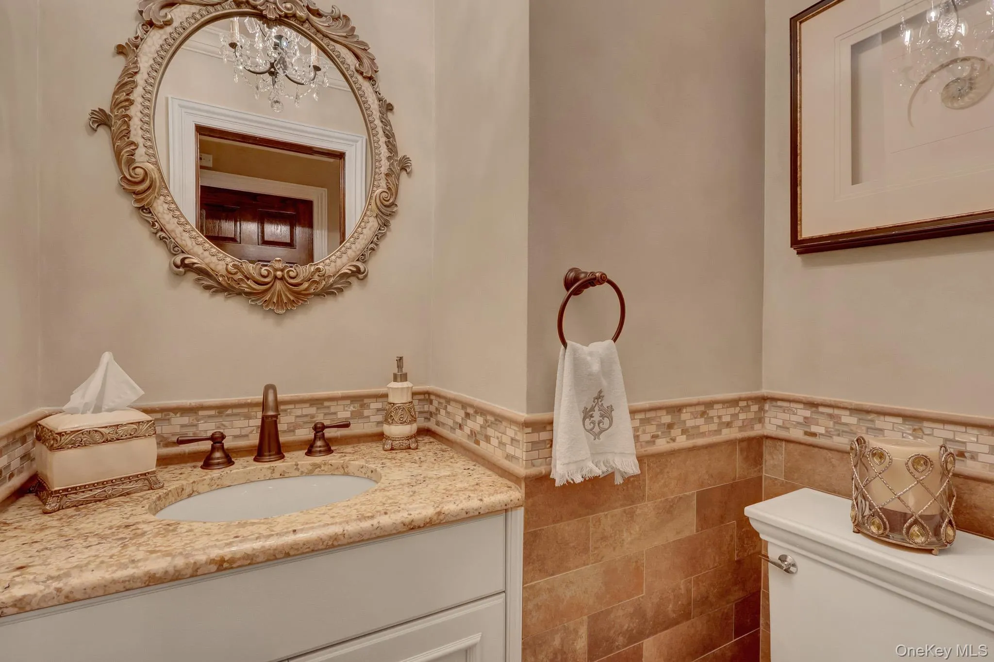 Powder room featuring a decorative framed mirror, a vanity with a stone countertop, a bronze-finish faucet, and wainscoting with mosaic tile accents Powder room featuring a decorative framed mirror, a vanity with a stone countertop, a bronze-finish faucet, and wainscoting with mosaic tile accents