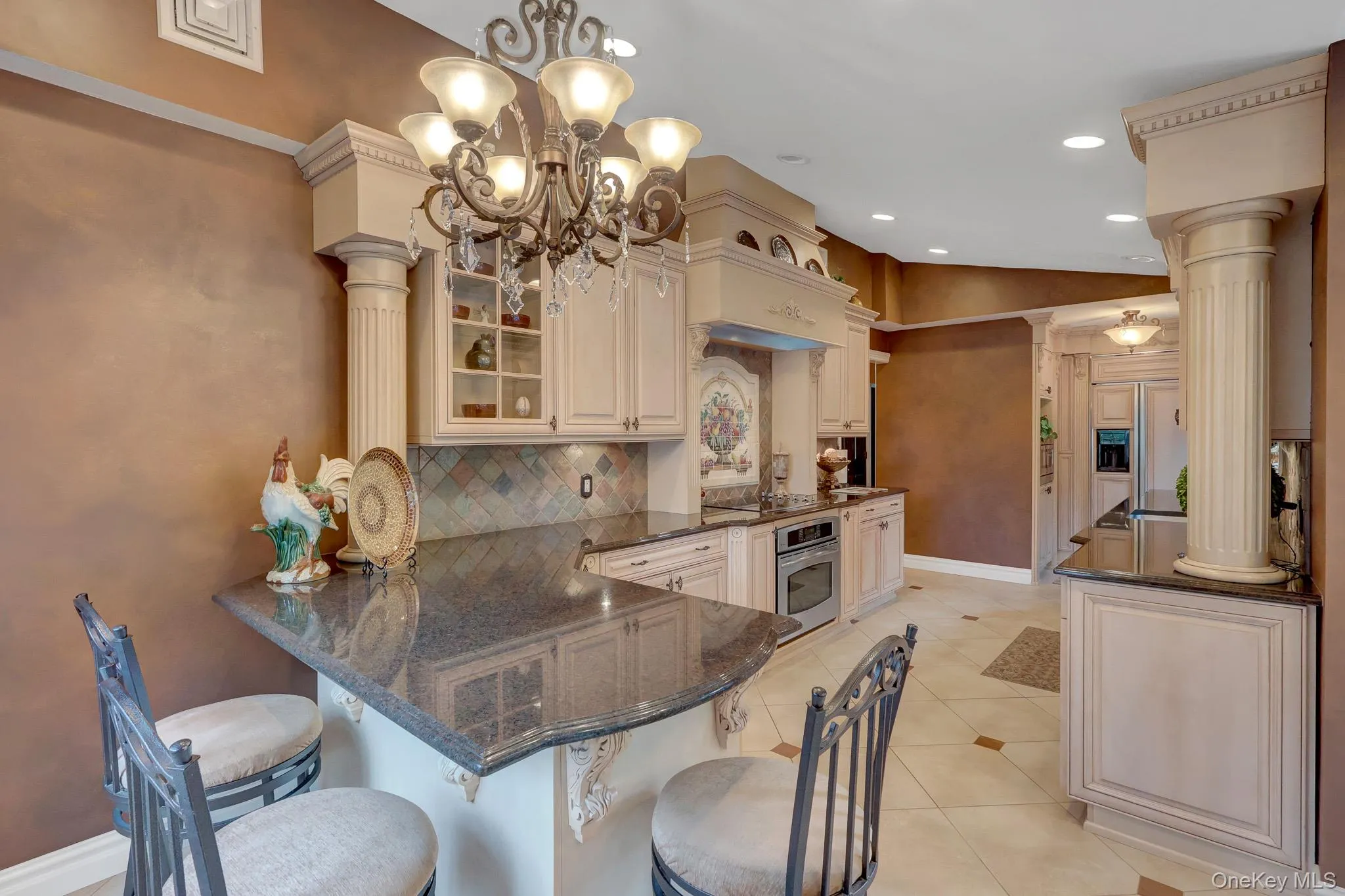 Gourmet kitchen featuring cream-colored cabinetry, dark countertops, built-in oven, detailed backsplash, and a decorative chandelier Gourmet kitchen featuring cream-colored cabinetry, dark countertops, built-in oven, detailed backsplash, and a decorative chandelier