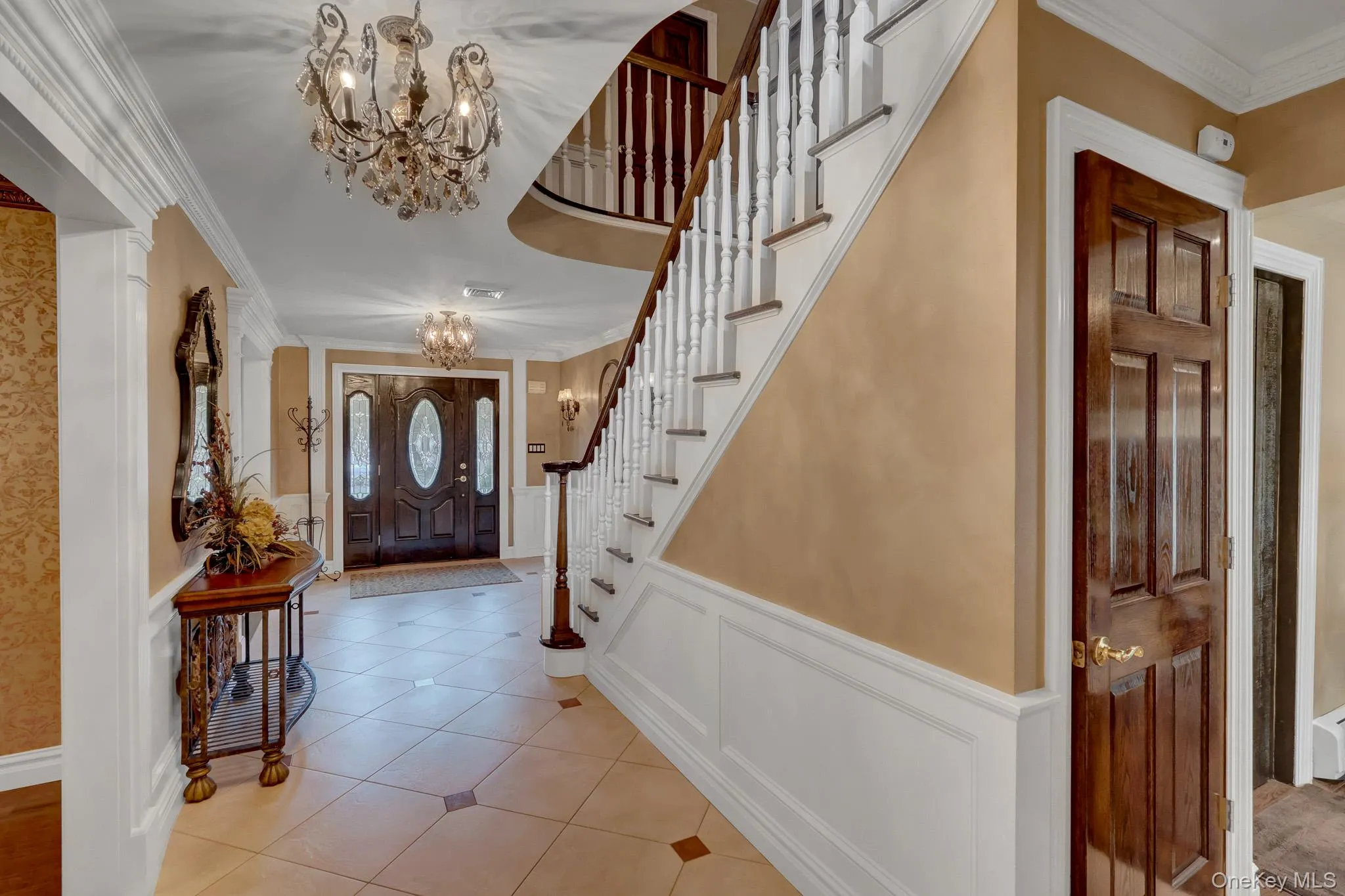 Grand entryway featuring a decorative glass-paneled double door, a curved staircase with white balusters and dark wood handrail, and tile flooring with accent squares Grand entryway featuring a decorative glass-paneled double door, a curved staircase with white balusters and dark wood handrail, and tile flooring with accent squares