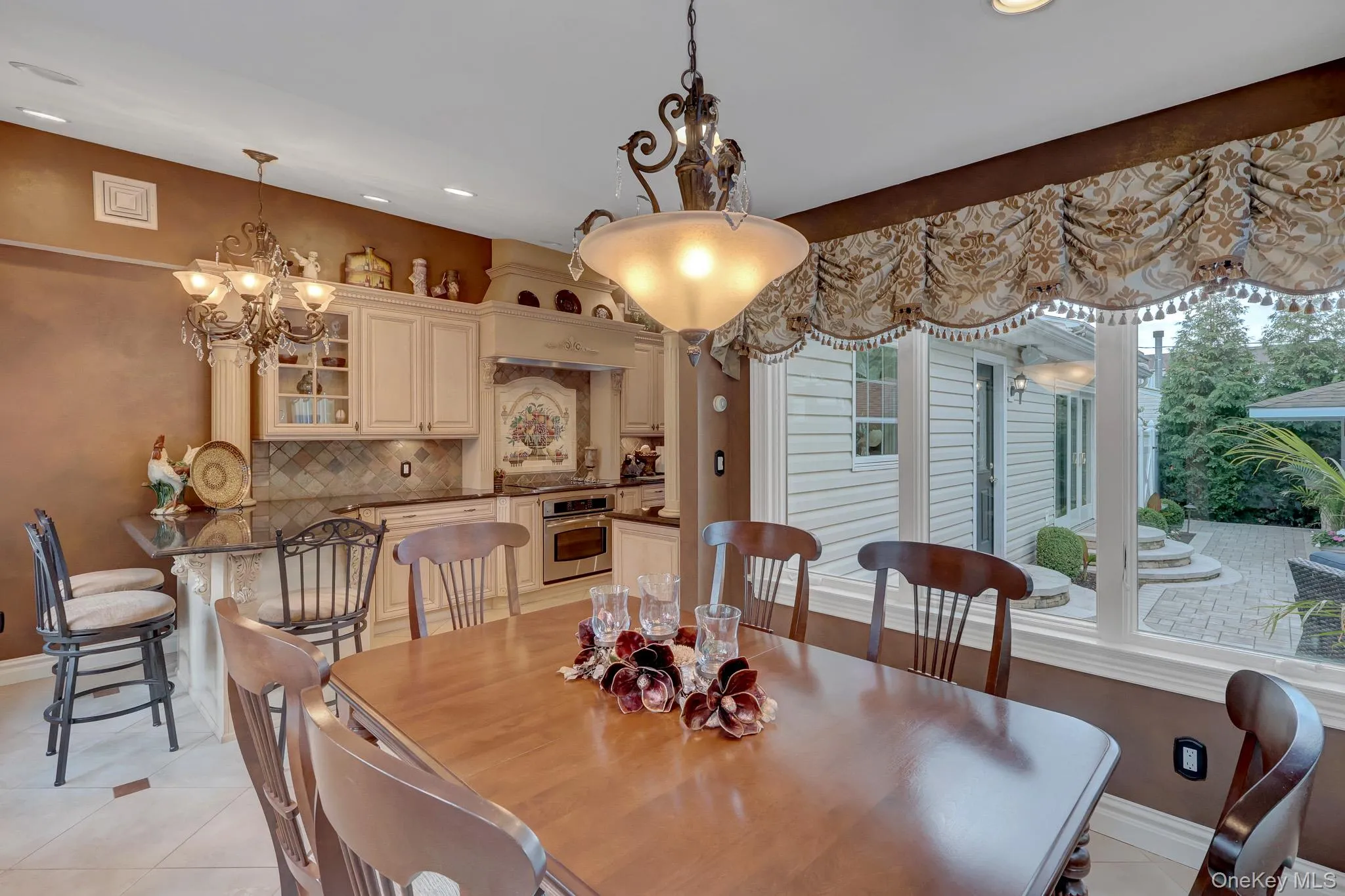 Dining area with wood-finish flooring, featuring a classic chandelier and a large window overlooking a paved patio Dining area with wood-finish flooring, featuring a classic chandelier and a large window overlooking a paved patio