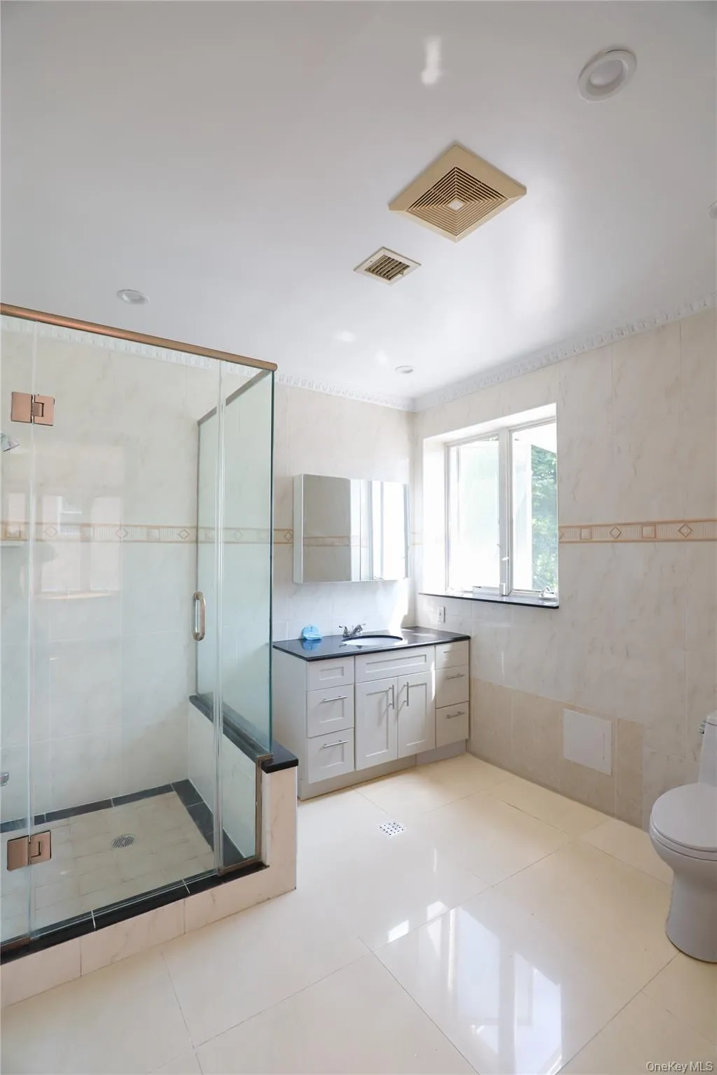 Bathroom featuring a glass-enclosed shower with tile surround, a single vanity with a dark countertop and undermount sink, a toilet, and large format floor tiles Bathroom featuring a glass-enclosed shower with tile surround, a single vanity with a dark countertop and undermount sink, a toilet, and large format floor tiles
