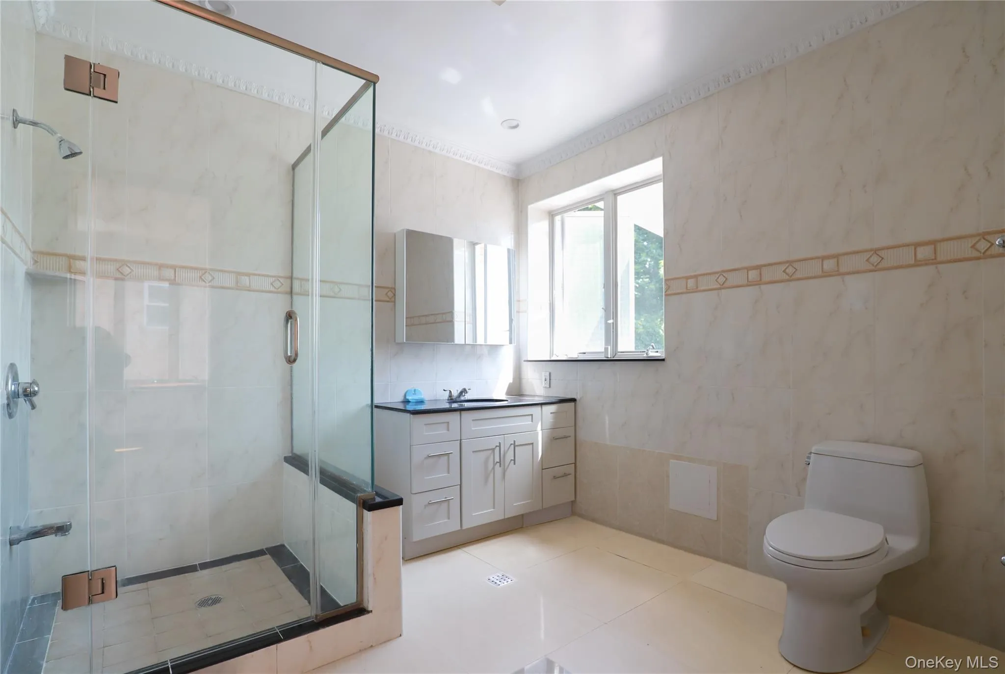 Tiled bathroom featuring a glass-enclosed shower, a vanity with an undermount sink and dark countertop, and a window for natural light Tiled bathroom featuring a glass-enclosed shower, a vanity with an undermount sink and dark countertop, and a window for natural light
