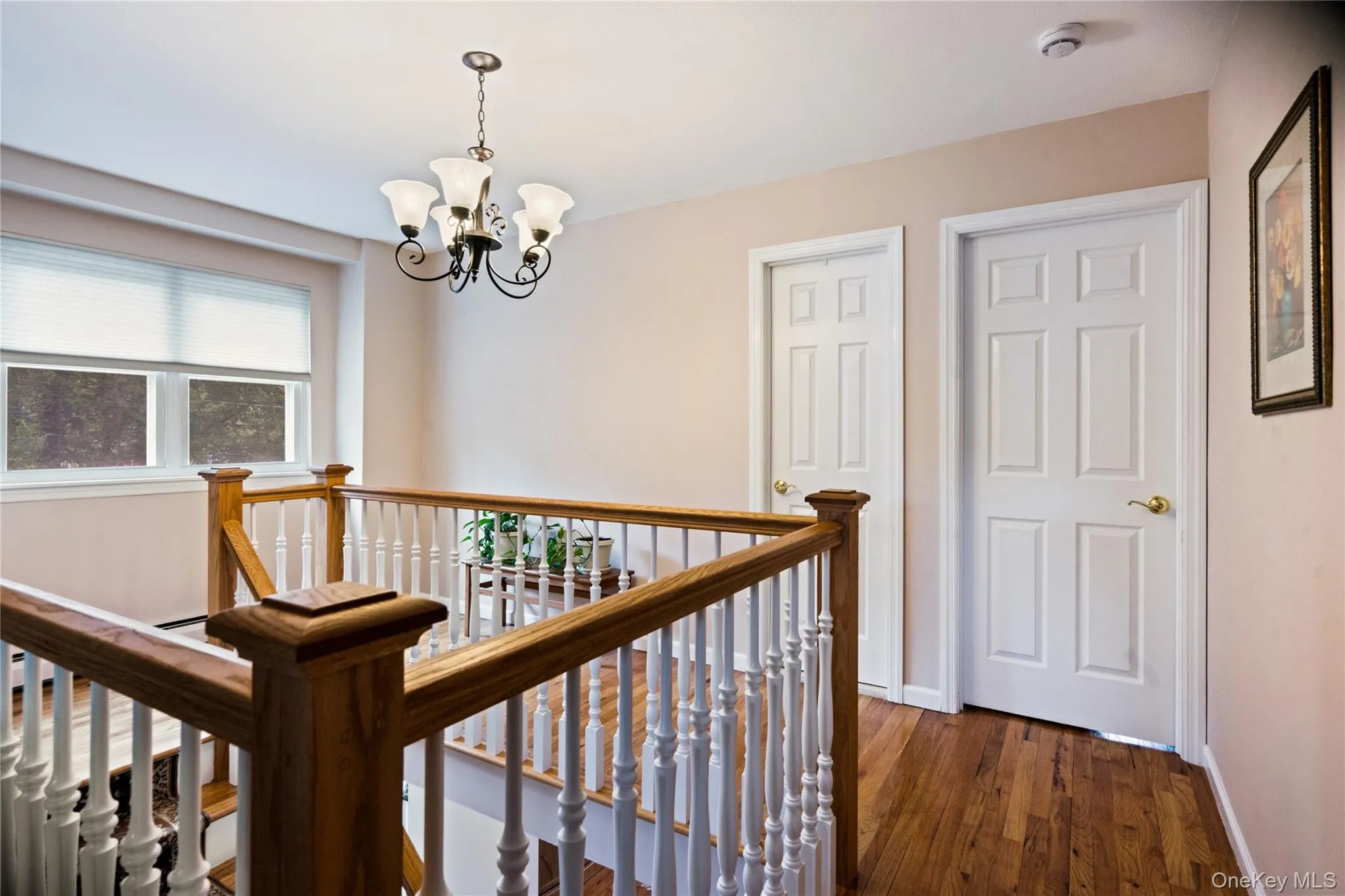 Staircase landing featuring wood-finish flooring, a traditional chandelier, two paneled doors with brass hardware, a light wood-finish banister, and a window with blinds Staircase landing featuring wood-finish flooring, a traditional chandelier, two paneled doors with brass hardware, a light wood-finish banister, and a window with blinds