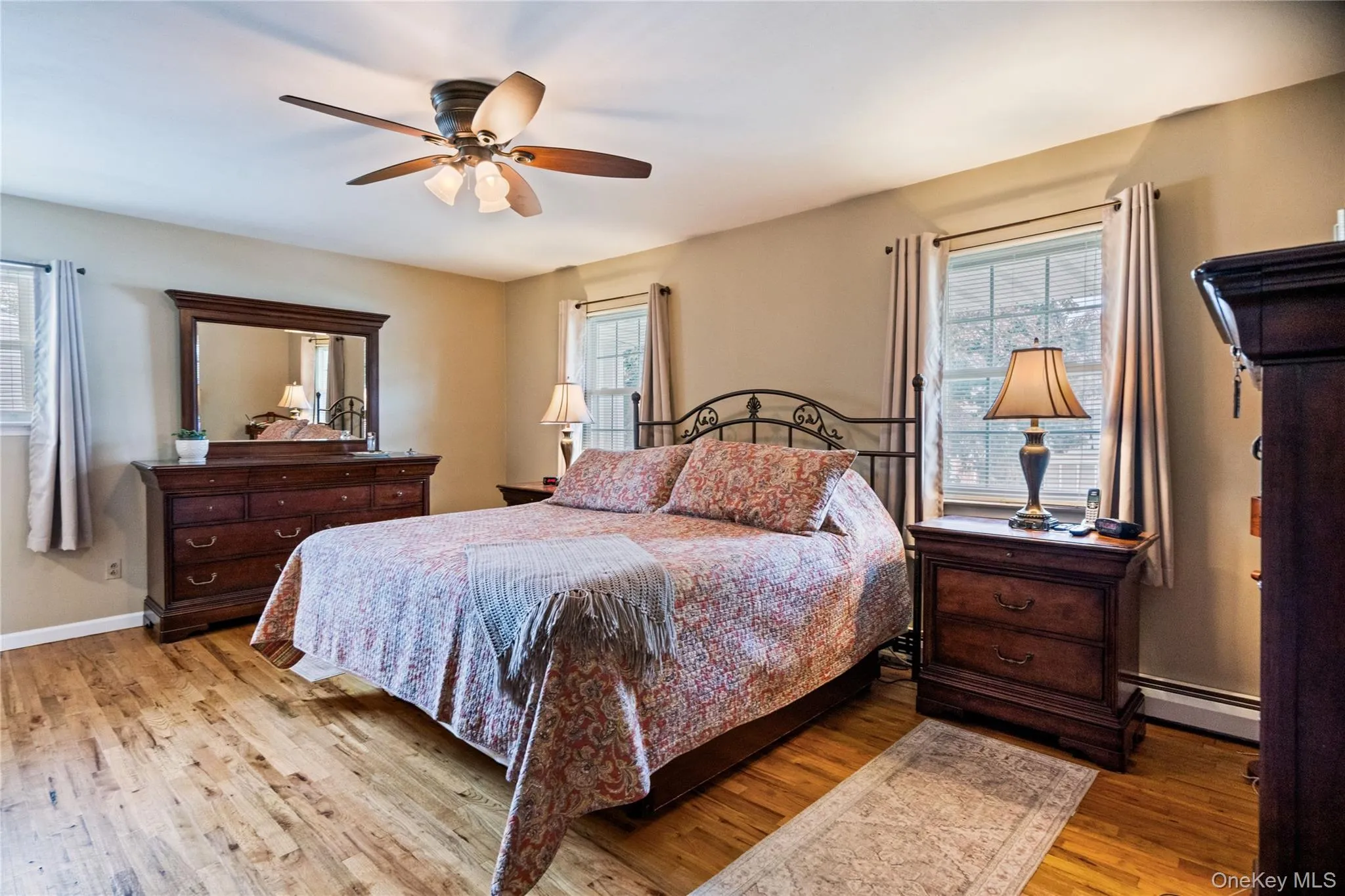 Room featuring wood-finish flooring, two windows with blinds, a ceiling fan with light fixture, and neutral-toned wall paint Room featuring wood-finish flooring, two windows with blinds, a ceiling fan with light fixture, and neutral-toned wall paint