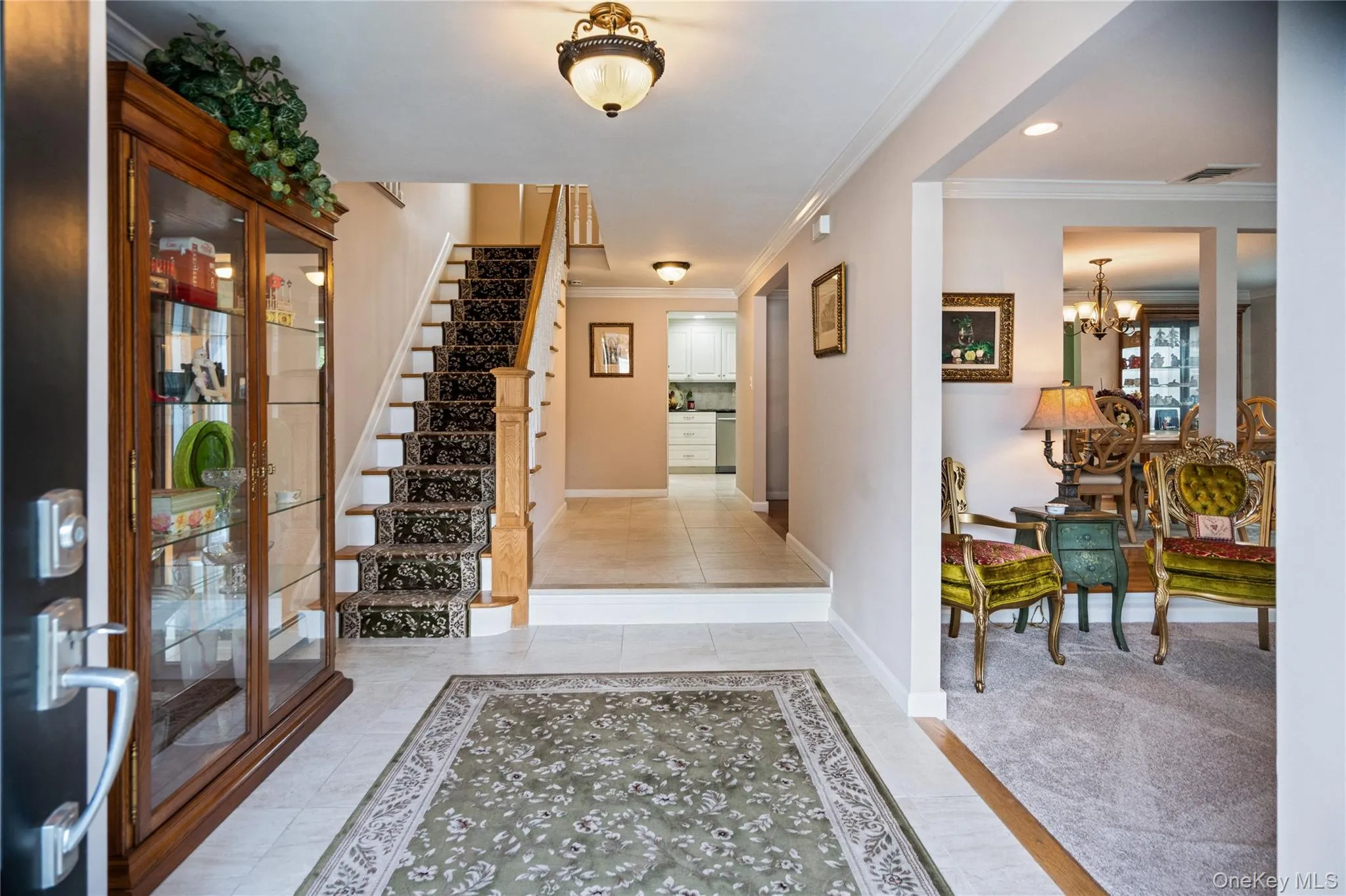 Tiled entryway with decorative area rug, leading to a carpeted staircase with wood banister Tiled entryway with decorative area rug, leading to a carpeted staircase with wood banister