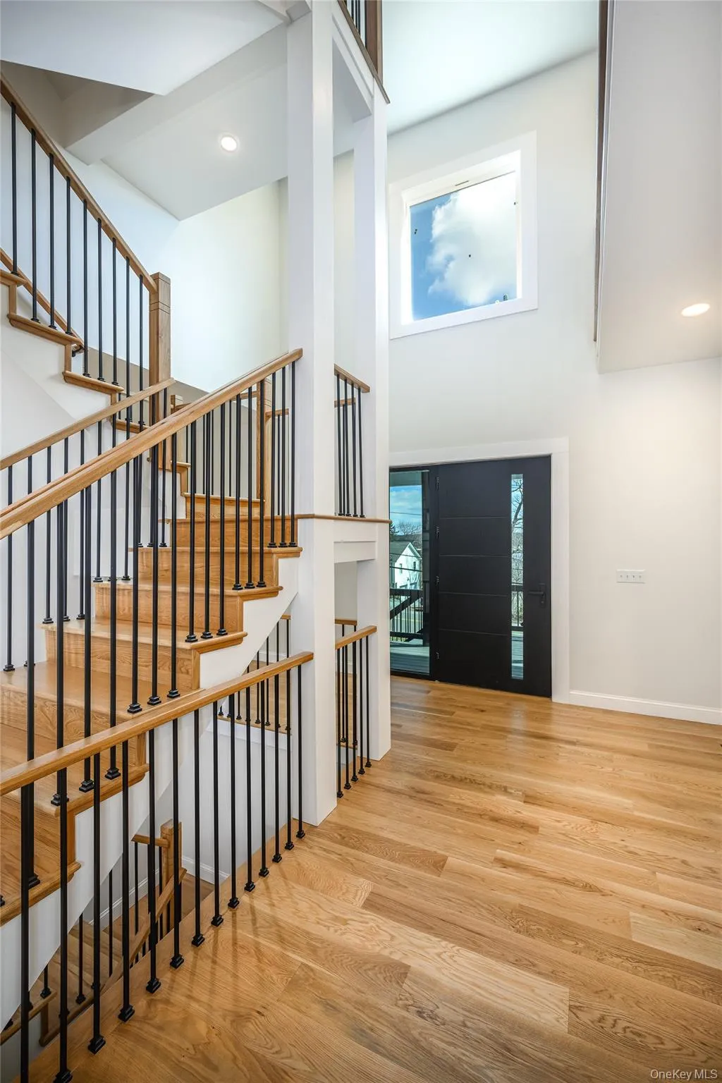Two-story foyer featuring wood-finish flooring, a grand staircase with wood railings and black iron balusters, a tall window, recessed lighting, and a modern dark-paneled entry door Two-story foyer featuring wood-finish flooring, a grand staircase with wood railings and black iron balusters, a tall window, recessed lighting, and a modern dark-paneled entry door