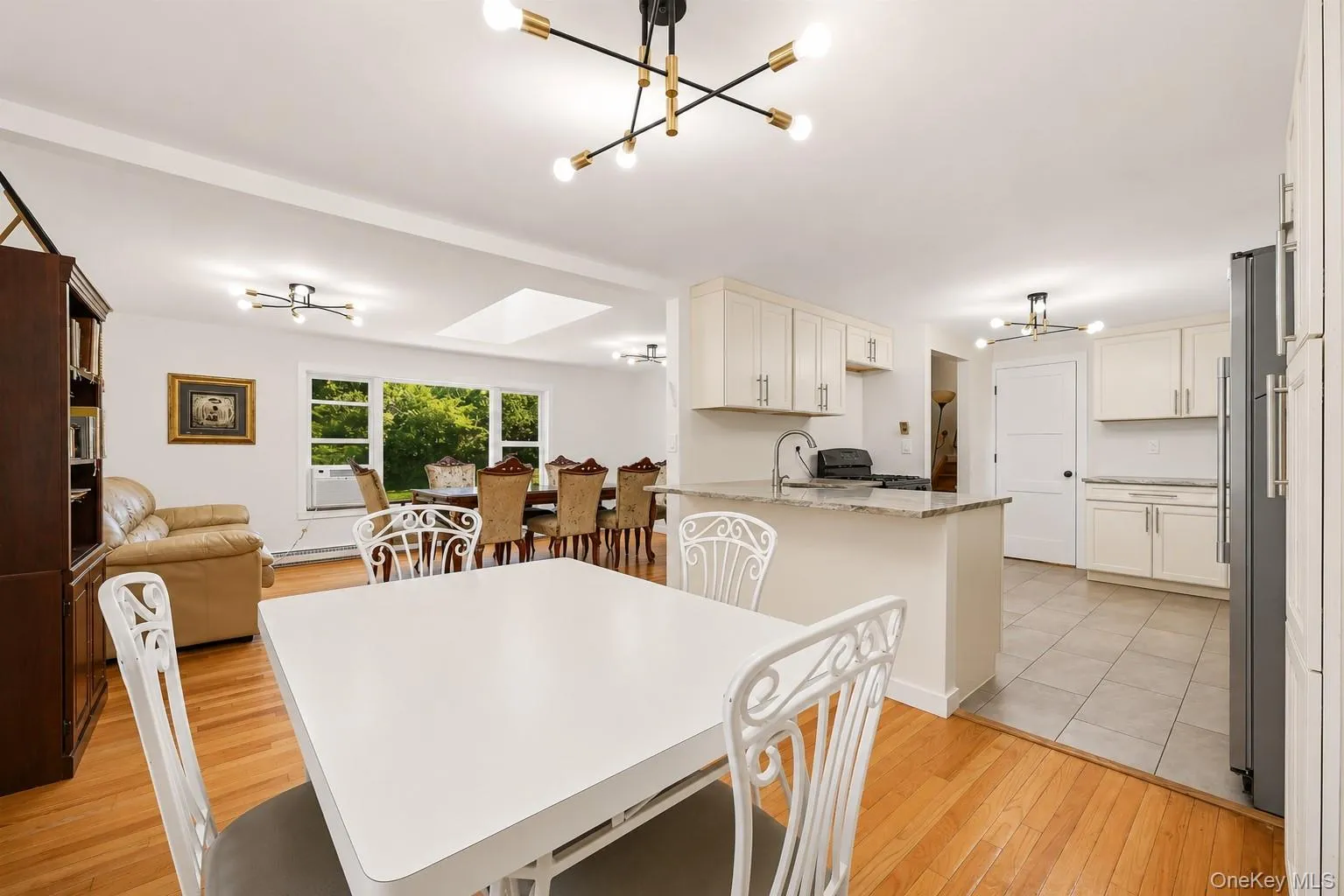 Open-concept space featuring hardwood flooring, a skylight, and a kitchen with light-toned cabinetry, a breakfast bar, and tile flooring Open-concept space featuring hardwood flooring, a skylight, and a kitchen with light-toned cabinetry, a breakfast bar, and tile flooring