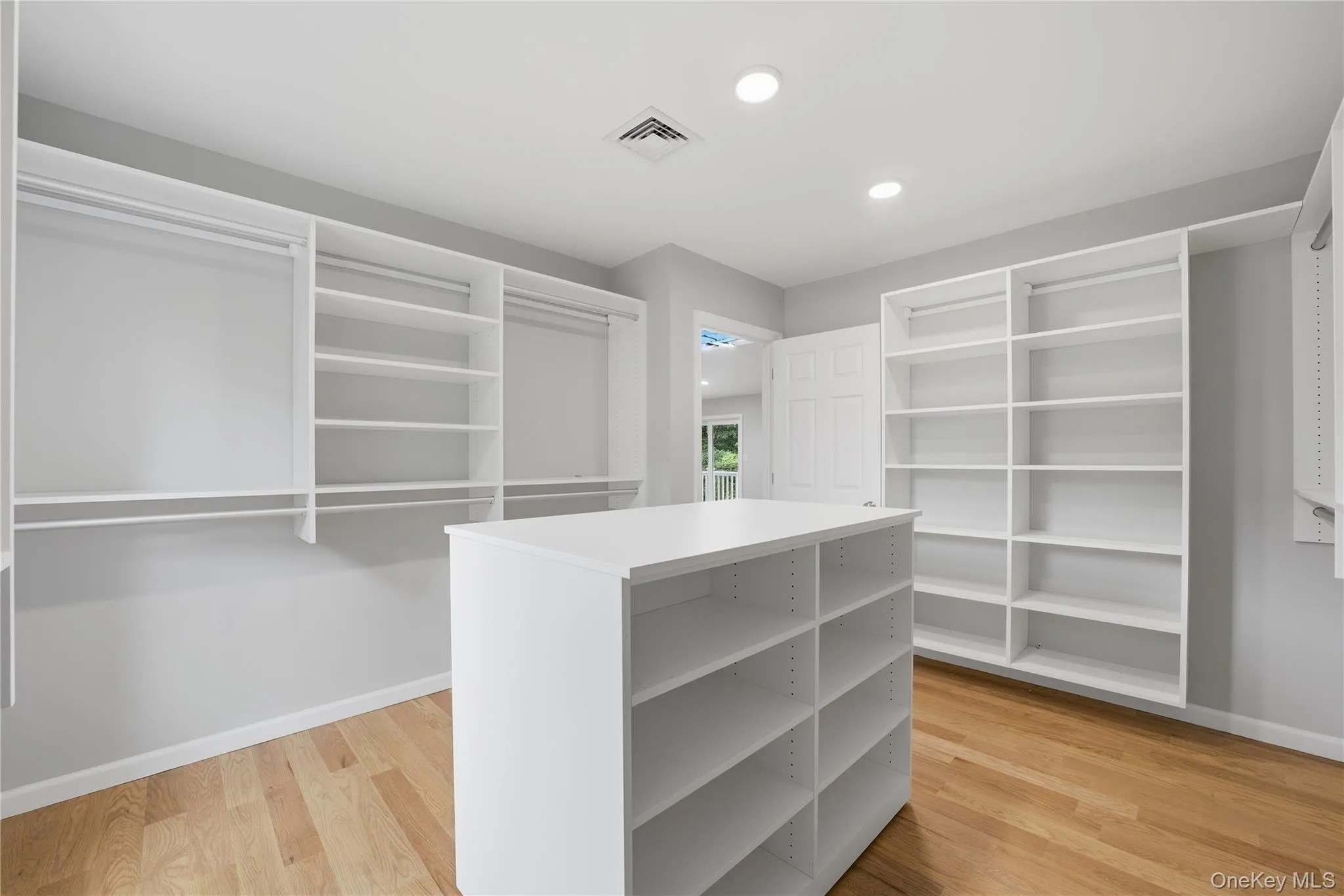 Spacious custom closet featuring extensive shelving, multiple hanging rods, a central storage island, recessed lighting, and wood-finish flooring Spacious custom closet featuring extensive shelving, multiple hanging rods, a central storage island, recessed lighting, and wood-finish flooring