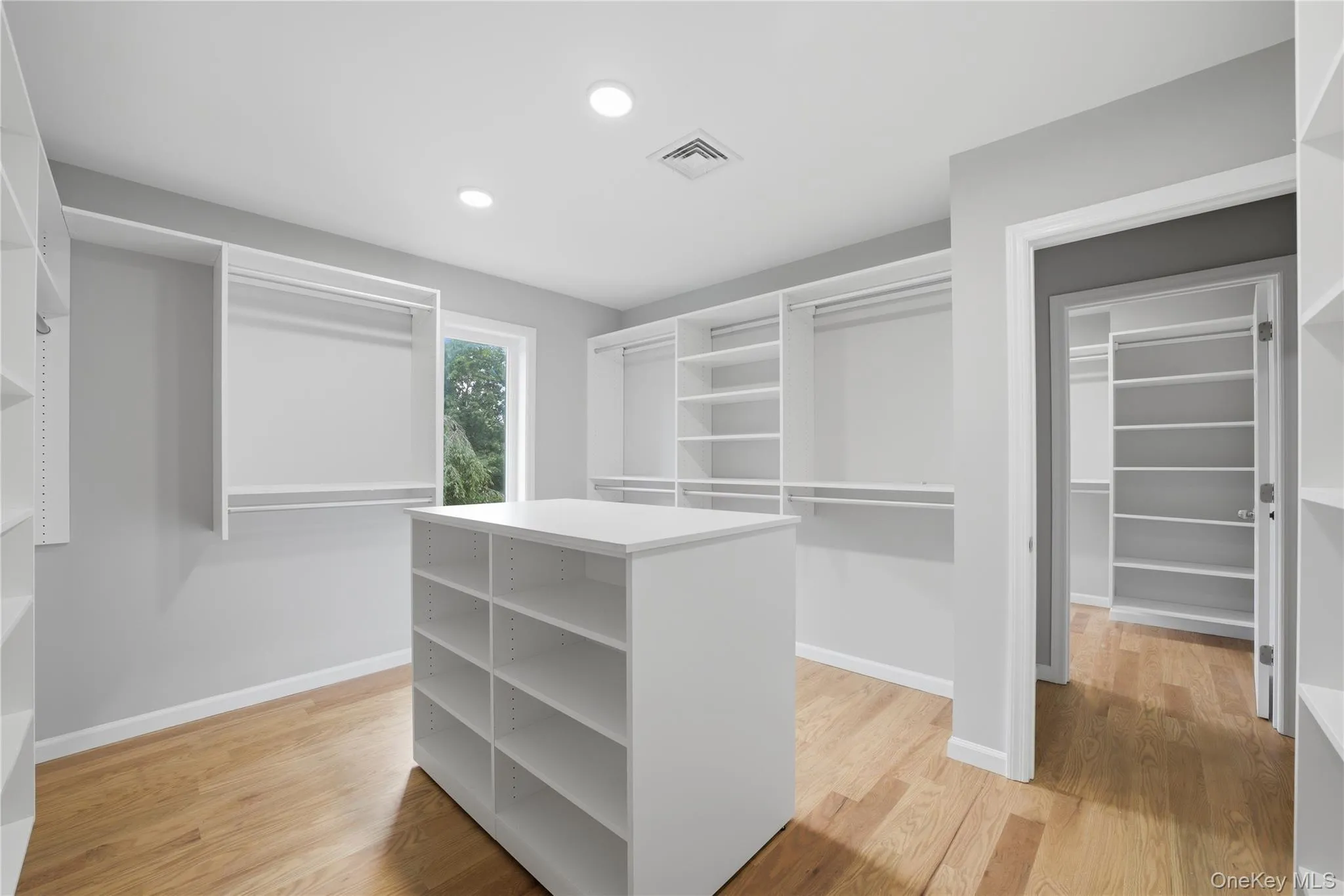 Spacious walk-in closet featuring extensive custom built-in shelving, hanging rods, a central storage island, wood-finish flooring, and recessed lighting Spacious walk-in closet featuring extensive custom built-in shelving, hanging rods, a central storage island, wood-finish flooring, and recessed lighting