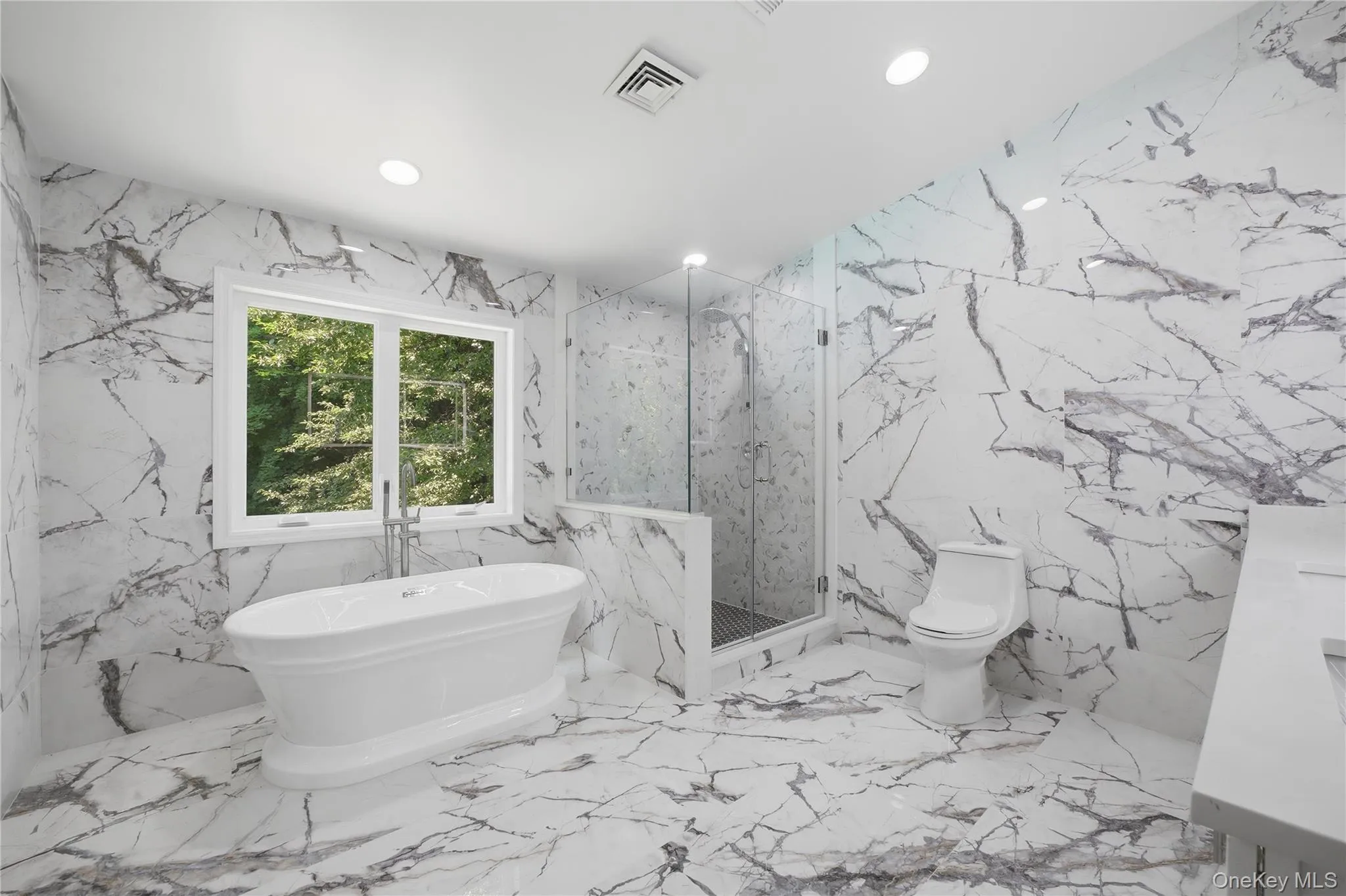 Spacious primary ensuite bathroom featuring large format marble-look tile flooring and walls, a freestanding soaking tub with floor-mounted faucet, a glass-enclosed shower, and a white vanity with integrated sink Spacious primary ensuite bathroom featuring large format marble-look tile flooring and walls, a freestanding soaking tub with floor-mounted faucet, a glass-enclosed shower, and a white vanity with integrated sink