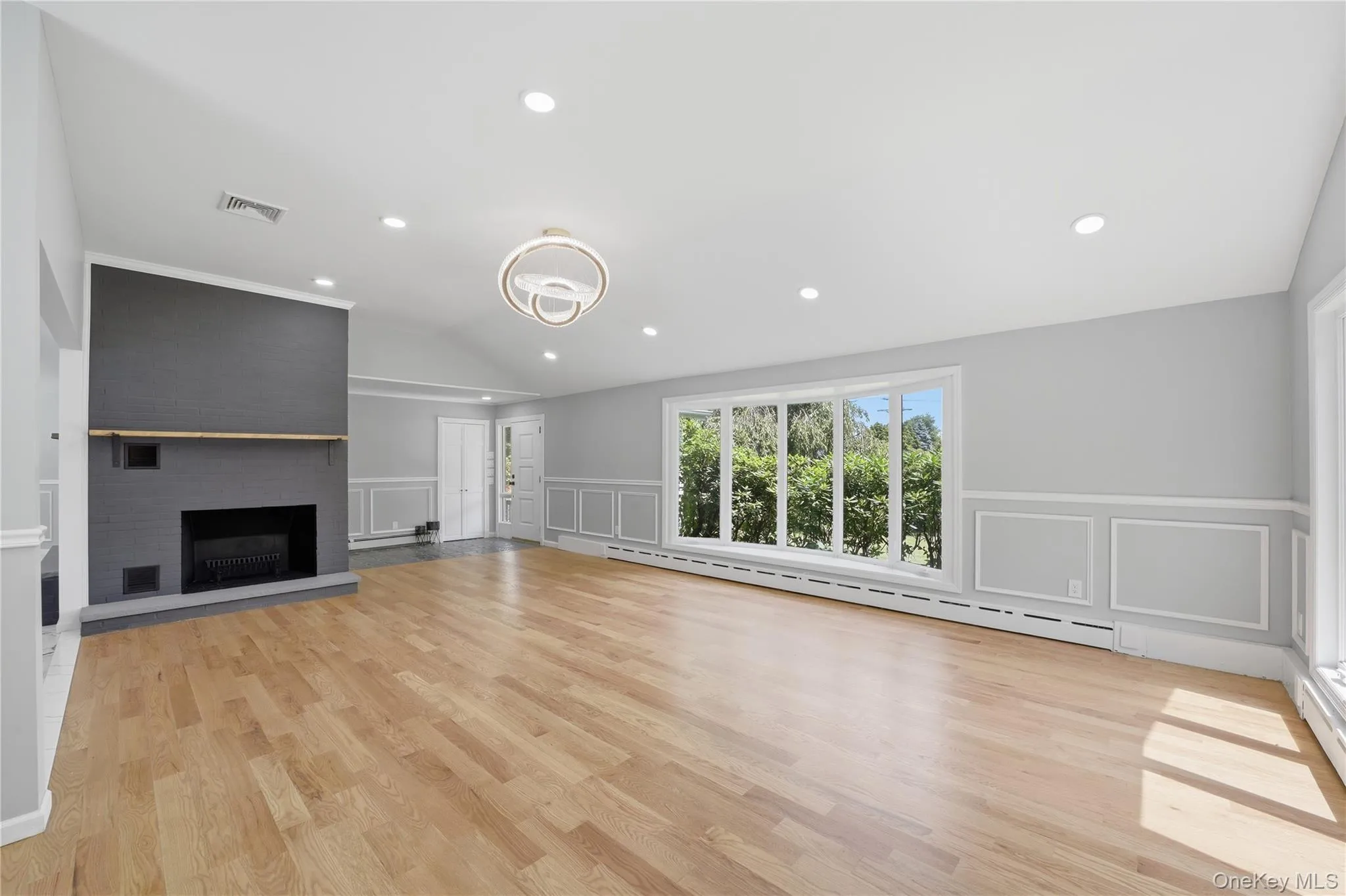 Spacious interior with natural wood flooring, a fireplace featuring a dark gray brick surround and wood mantel, a large bay window, and wainscoting with decorative trim Spacious interior with natural wood flooring, a fireplace featuring a dark gray brick surround and wood mantel, a large bay window, and wainscoting with decorative trim