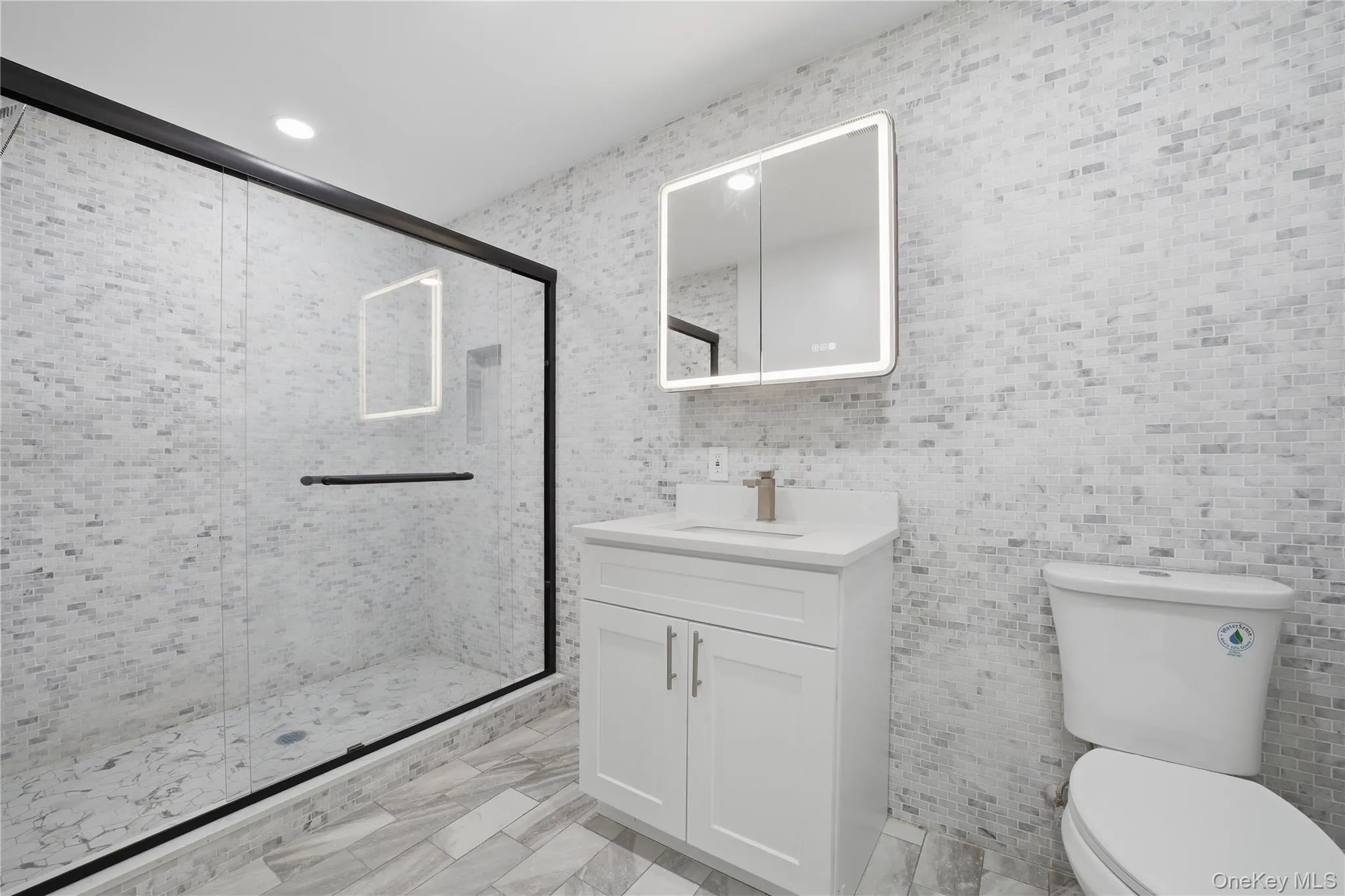 Main floor full modern bathroom featuring a frameless glass shower enclosure with matte black hardware, a white vanity with an integrated sink, an illuminated medicine cabinet, a standard toilet, and grey-toned tile flooring Main floor full modern bathroom featuring a frameless glass shower enclosure with matte black hardware, a white vanity with an integrated sink, an illuminated medicine cabinet, a standard toilet, and grey-toned tile flooring