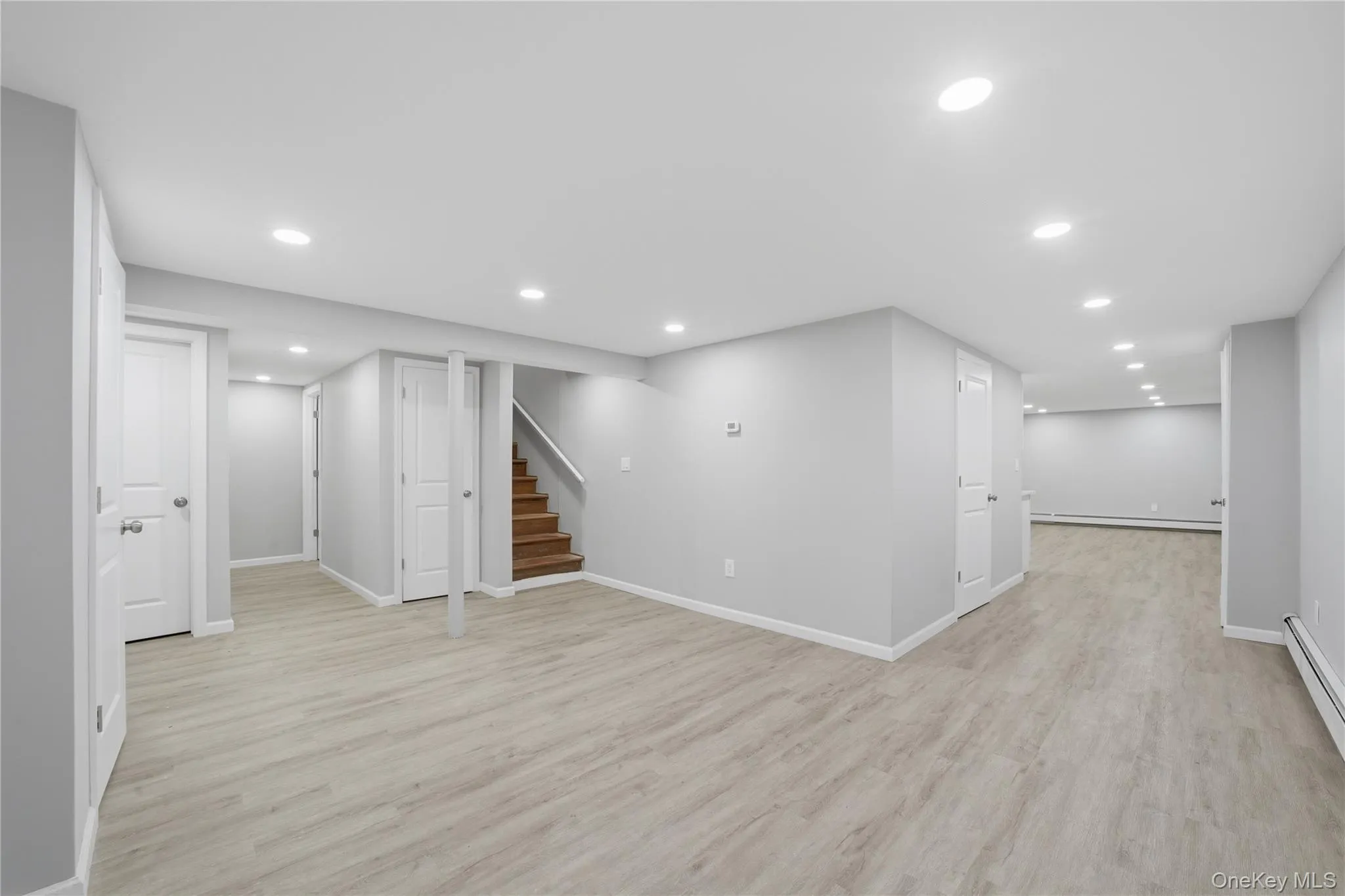 Spacious interior featuring light wood-finish flooring, recessed lighting, neutral wall tones, white trim, and a visible staircase with wood treads Spacious interior featuring light wood-finish flooring, recessed lighting, neutral wall tones, white trim, and a visible staircase with wood treads
