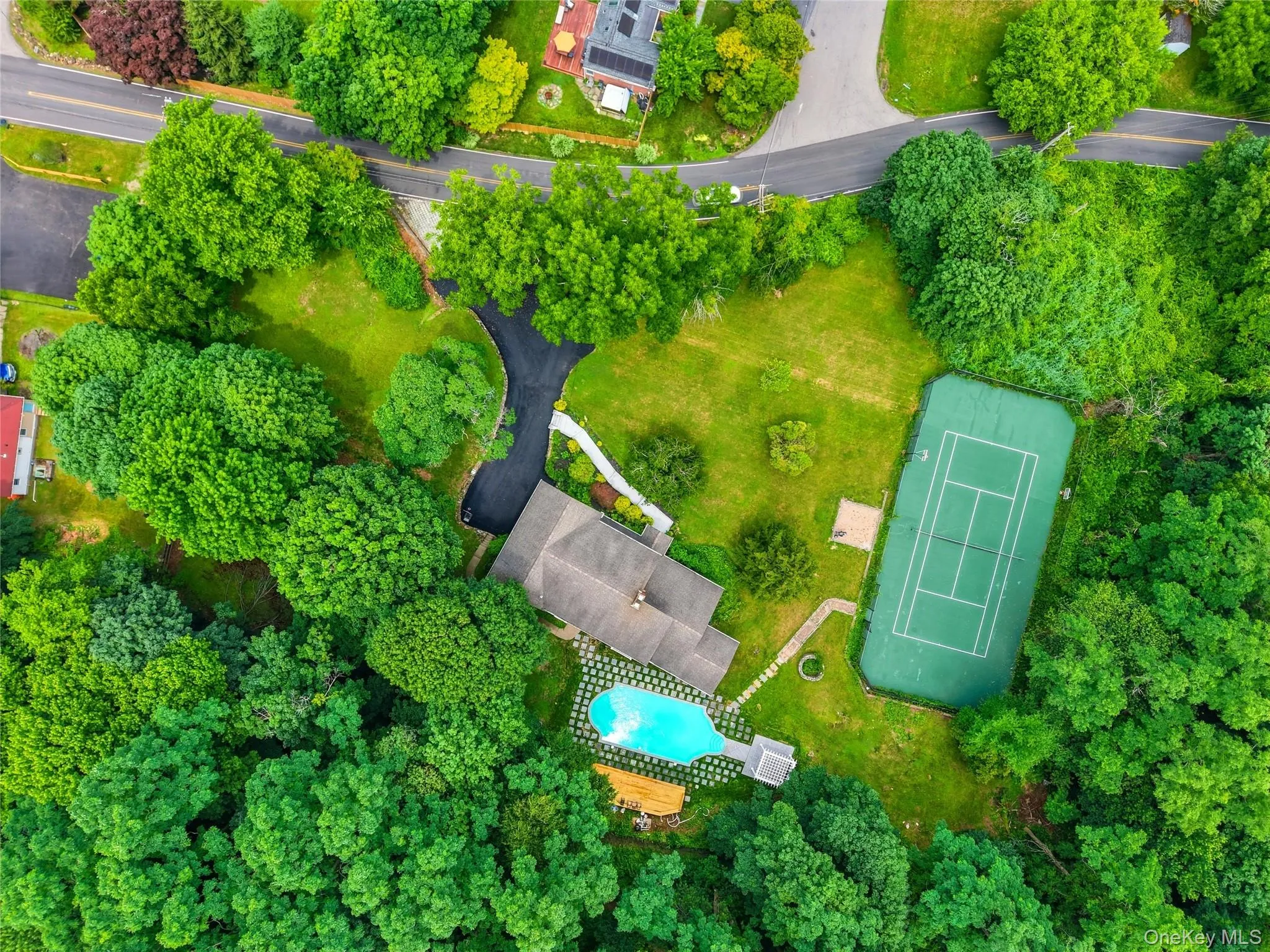 Expansive property featuring a private tennis court, a sparkling swimming pool, a substantial residence with a shingle roof, lush landscaping with mature trees, and a paved driveway Expansive property featuring a private tennis court, a sparkling swimming pool, a substantial residence with a shingle roof, lush landscaping with mature trees, and a paved driveway