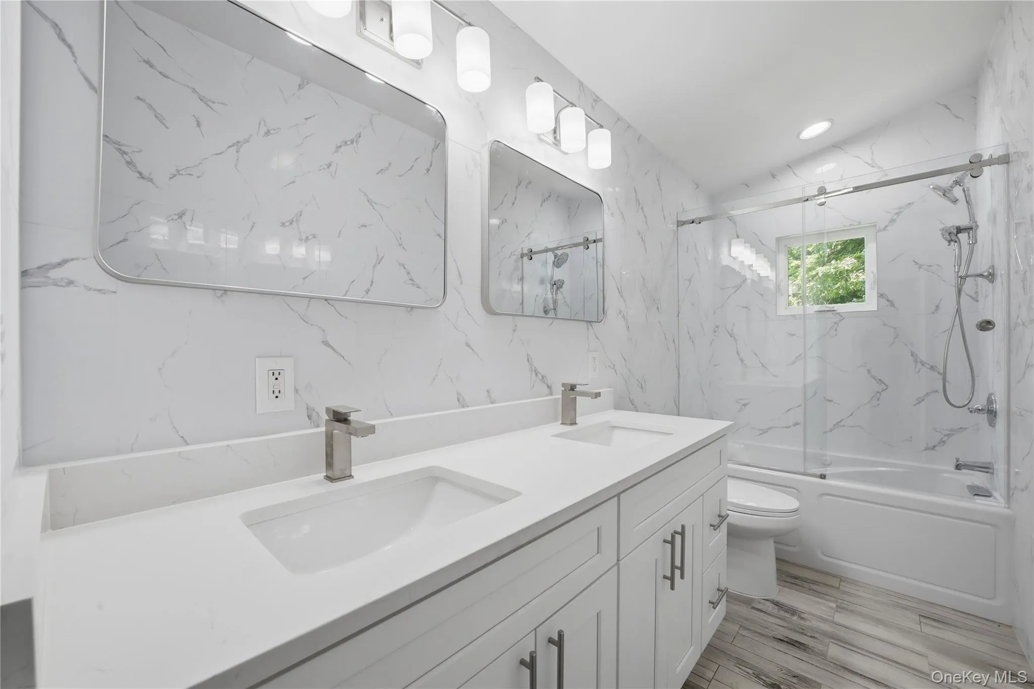 2nd floor full bathroom featuring a dual vanity with undermount sinks and brushed nickel faucets, a built-in bathtub with a sliding glass door, an overhead shower with a handheld attachment, and wood-finish flooring 2nd floor full bathroom featuring a dual vanity with undermount sinks and brushed nickel faucets, a built-in bathtub with a sliding glass door, an overhead shower with a handheld attachment, and wood-finish flooring