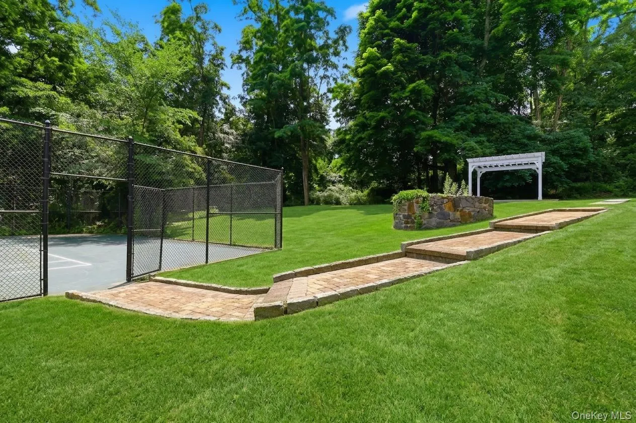 Spacious green lawn featuring a tennis court/ with black chain-link fencing, a multi-tiered paver pathway, a stone retaining wall, and a white pergola Spacious green lawn featuring a tennis court/ with black chain-link fencing, a multi-tiered paver pathway, a stone retaining wall, and a white pergola