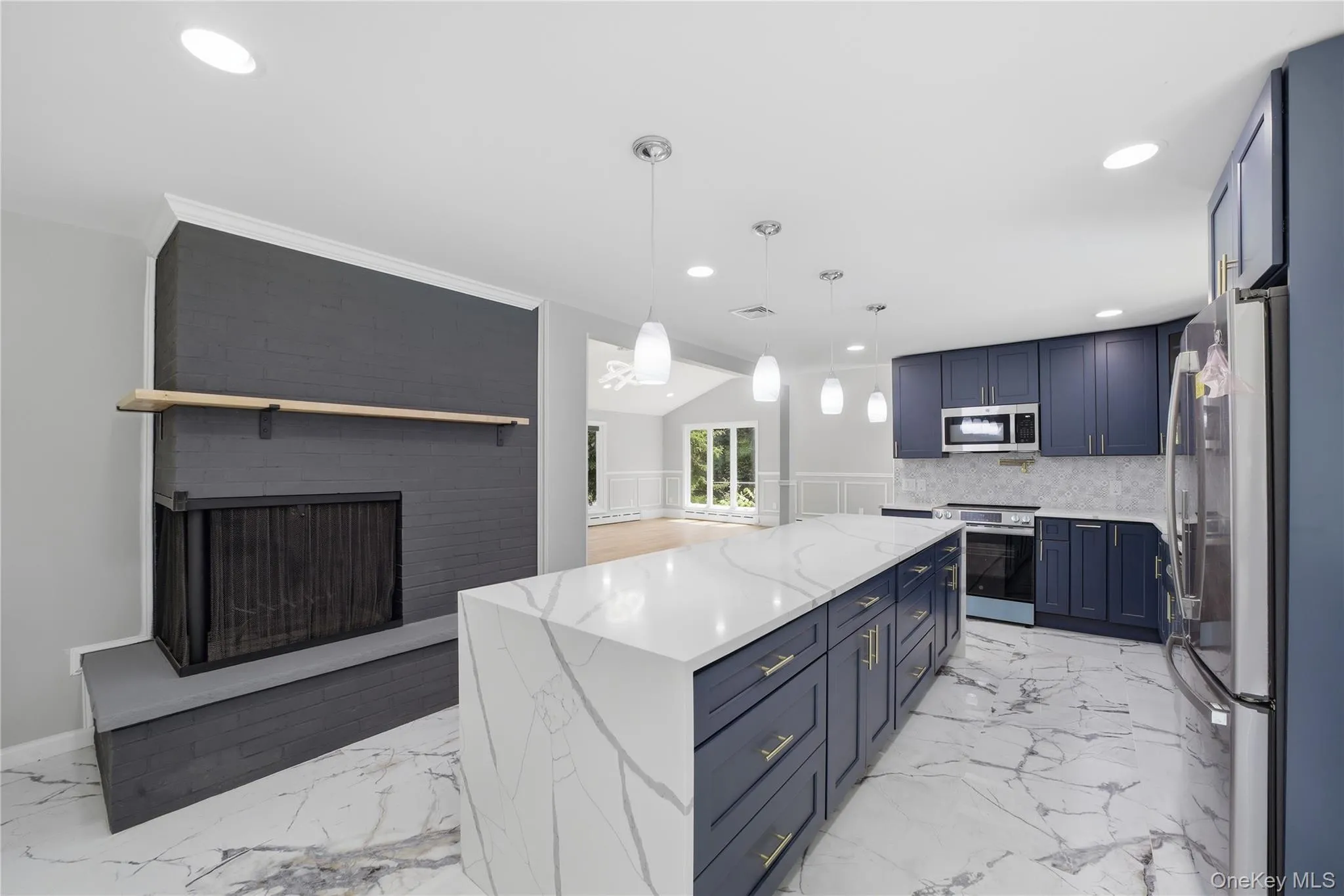 Open-concept kitchen featuring an expansive island with white veined countertop, custom navy cabinetry with gold-tone hardware, stainless steel appliances, and white marble-look flooring Open-concept kitchen featuring an expansive island with white veined countertop, custom navy cabinetry with gold-tone hardware, stainless steel appliances, and white marble-look flooring