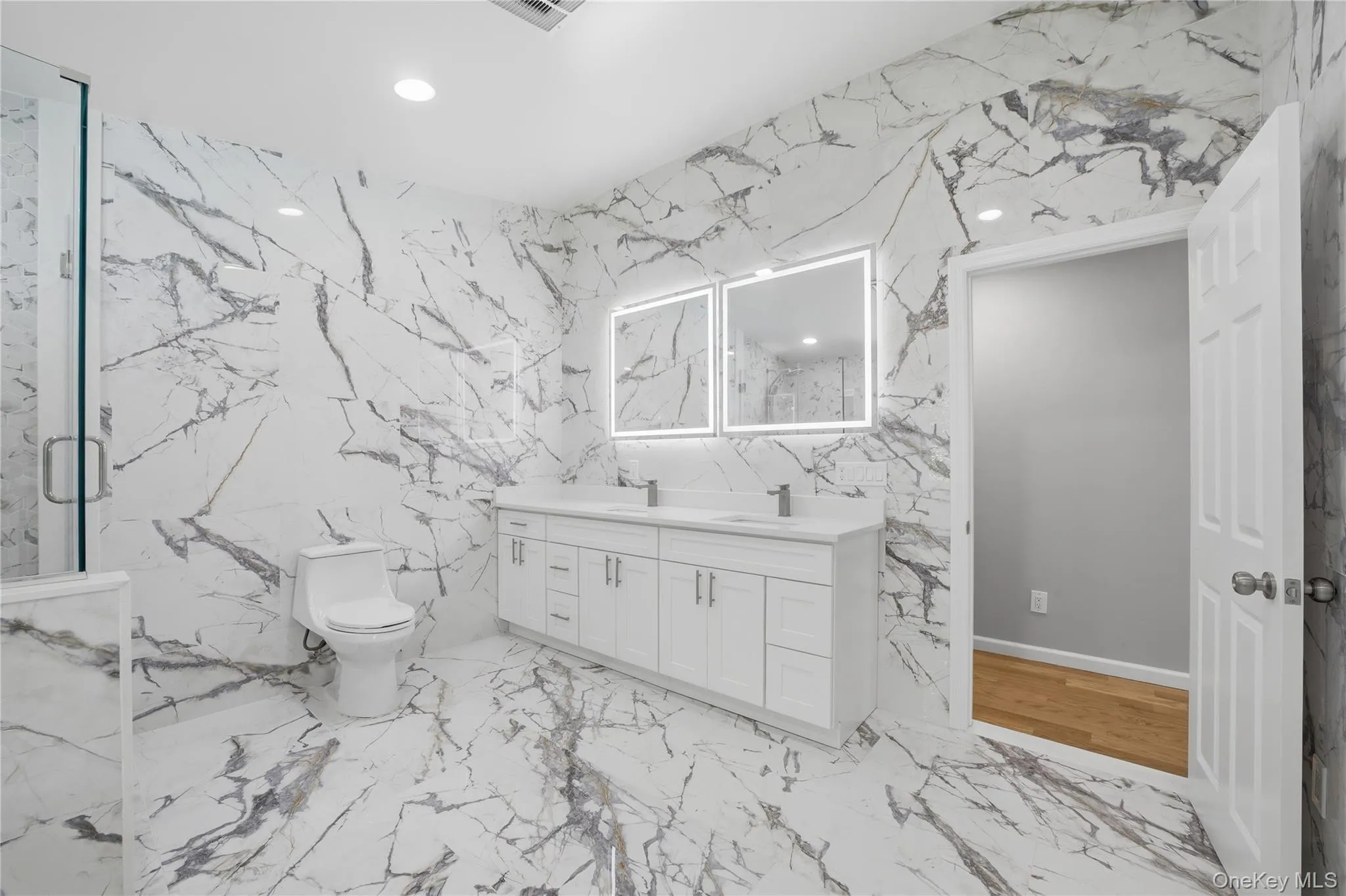 Primary ensuite bathroom featuring floor-to-ceiling white marble-look tile, a dual vanity with white cabinetry and integrated sinks, illuminated mirrors, and a glass-enclosed shower Primary ensuite bathroom featuring floor-to-ceiling white marble-look tile, a dual vanity with white cabinetry and integrated sinks, illuminated mirrors, and a glass-enclosed shower