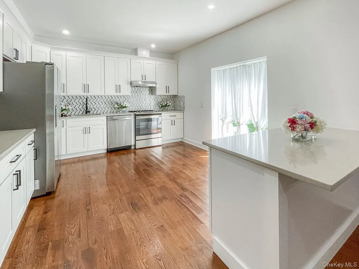 Modern kitchen featuring white shaker cabinetry, stainless steel appliances, a herringbone tile backsplash, light-toned countertops, and wood flooring Modern kitchen featuring white shaker cabinetry, stainless steel appliances, a herringbone tile backsplash, light-toned countertops, and wood flooring