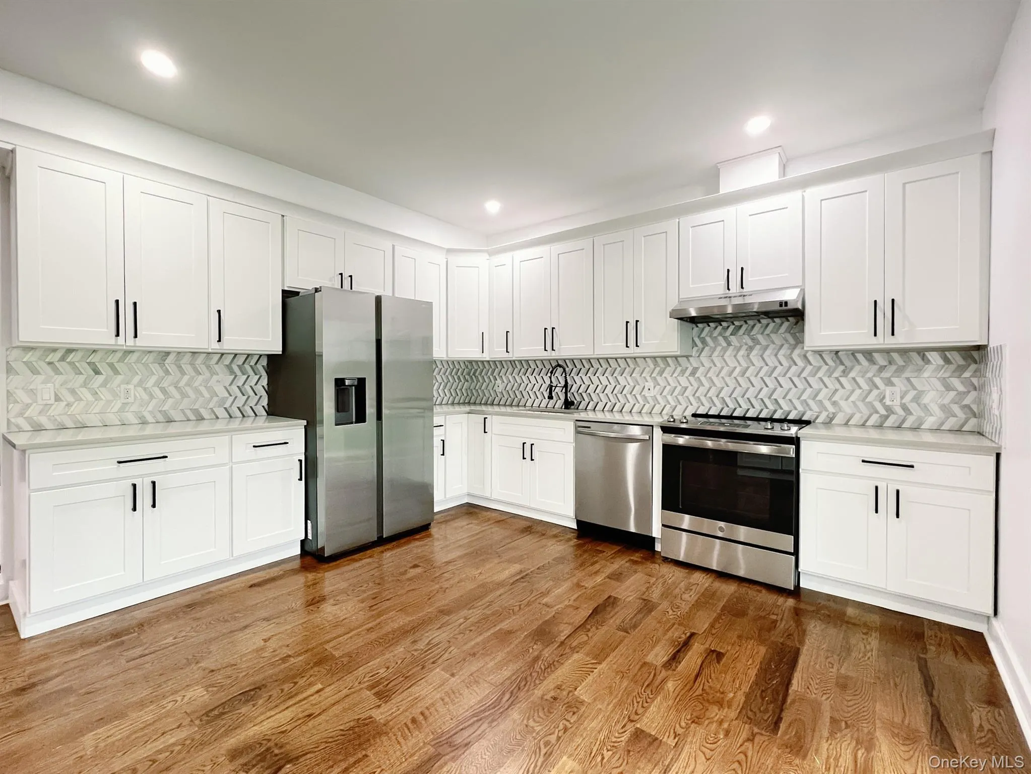 Modern kitchen featuring white shaker cabinetry, stainless steel appliances, herringbone tile backsplash, light countertops, and wood flooring Modern kitchen featuring white shaker cabinetry, stainless steel appliances, herringbone tile backsplash, light countertops, and wood flooring