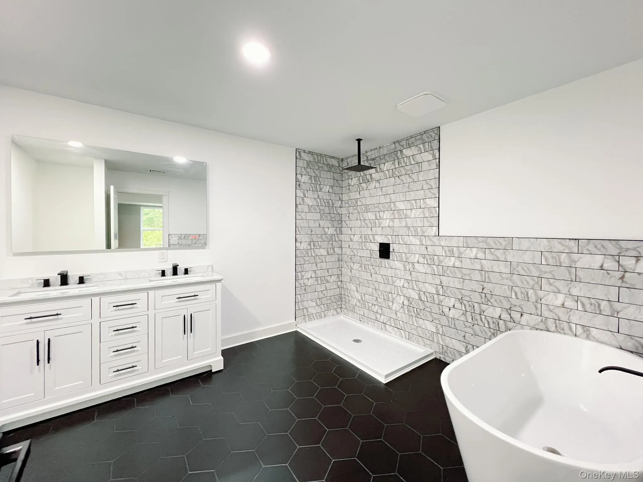 Spacious ensuite bathroom featuring a dual vanity with white cabinetry and dark hardware, marble-look countertop, large mirror, and a freestanding soaking tub Spacious ensuite bathroom featuring a dual vanity with white cabinetry and dark hardware, marble-look countertop, large mirror, and a freestanding soaking tub