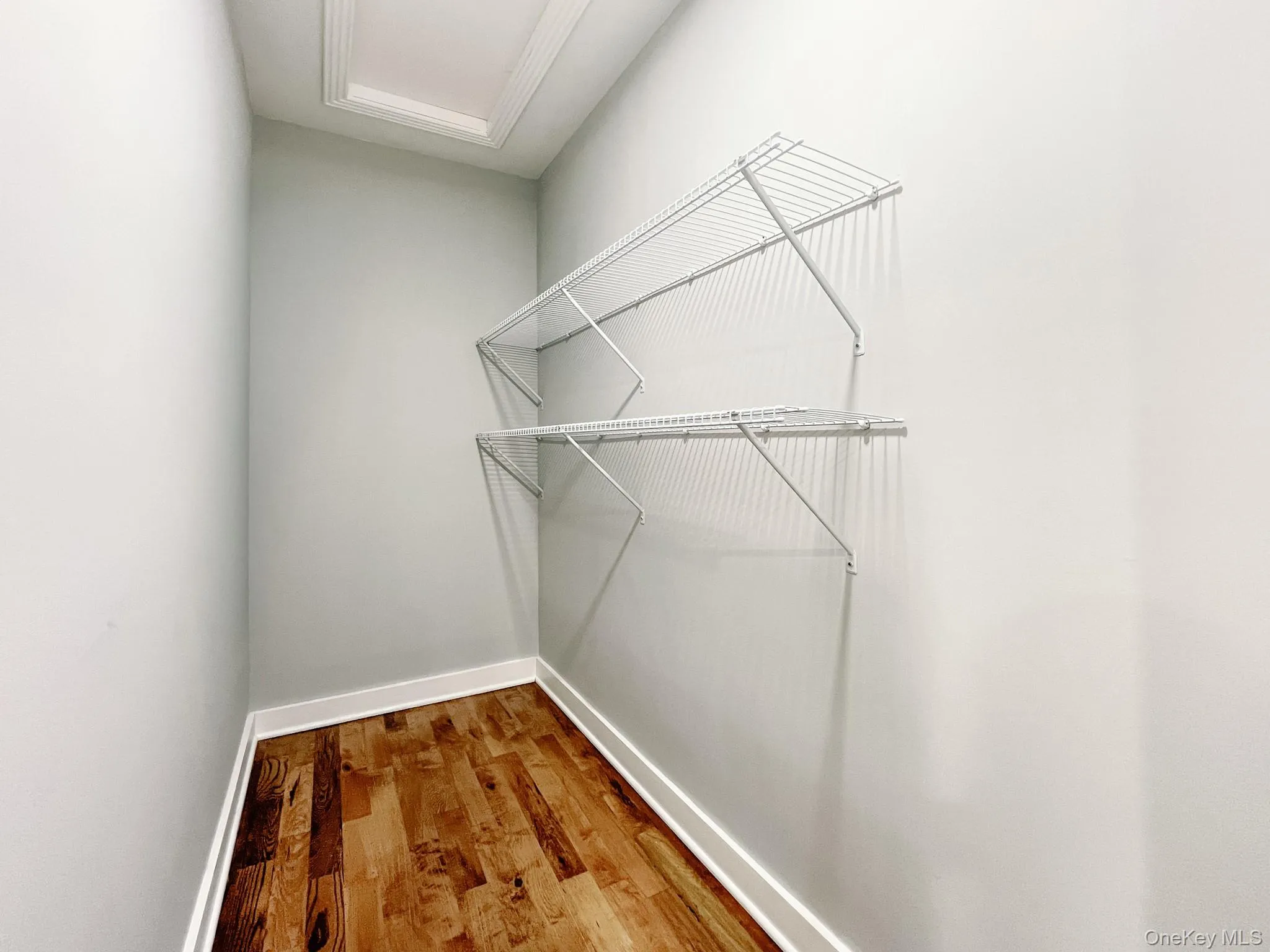 Bedroom #2 walk-in closet featuring two levels of wire shelving, wood-finish flooring, light grey wall paint, white baseboards, and a ceiling access panel Bedroom #2 walk-in closet featuring two levels of wire shelving, wood-finish flooring, light grey wall paint, white baseboards, and a ceiling access panel