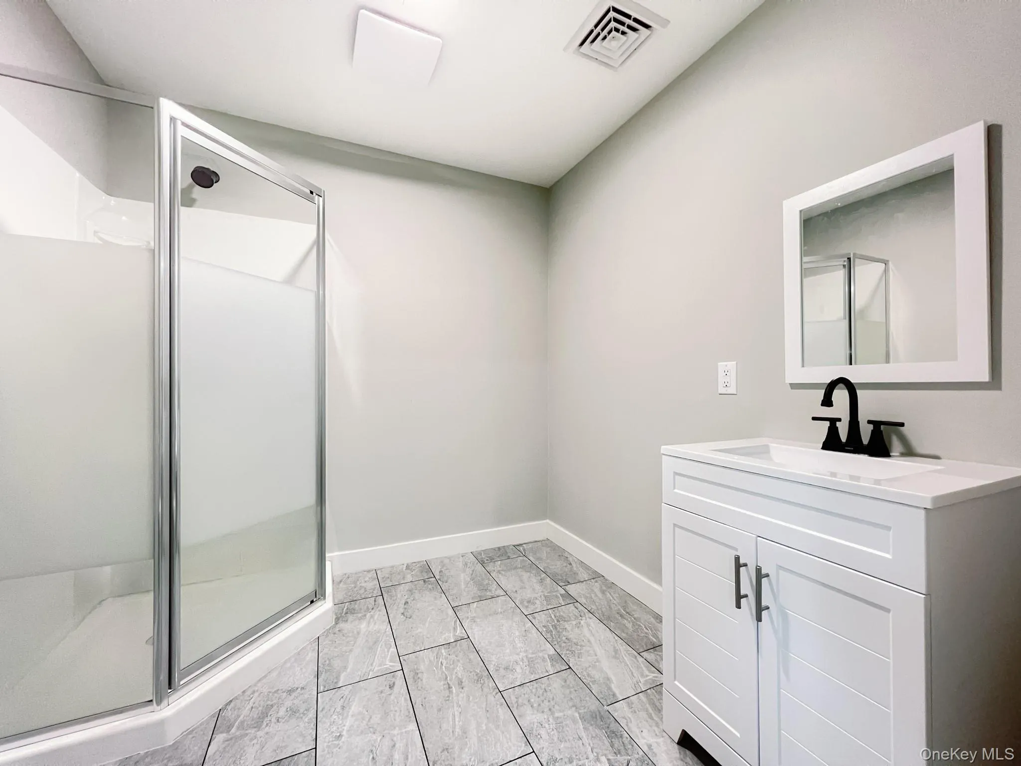 Ensuite bathroom featuring a glass-enclosed shower, white vanity with integrated sink, matte black faucet, framed mirror, and rectangular floor tiles Ensuite bathroom featuring a glass-enclosed shower, white vanity with integrated sink, matte black faucet, framed mirror, and rectangular floor tiles