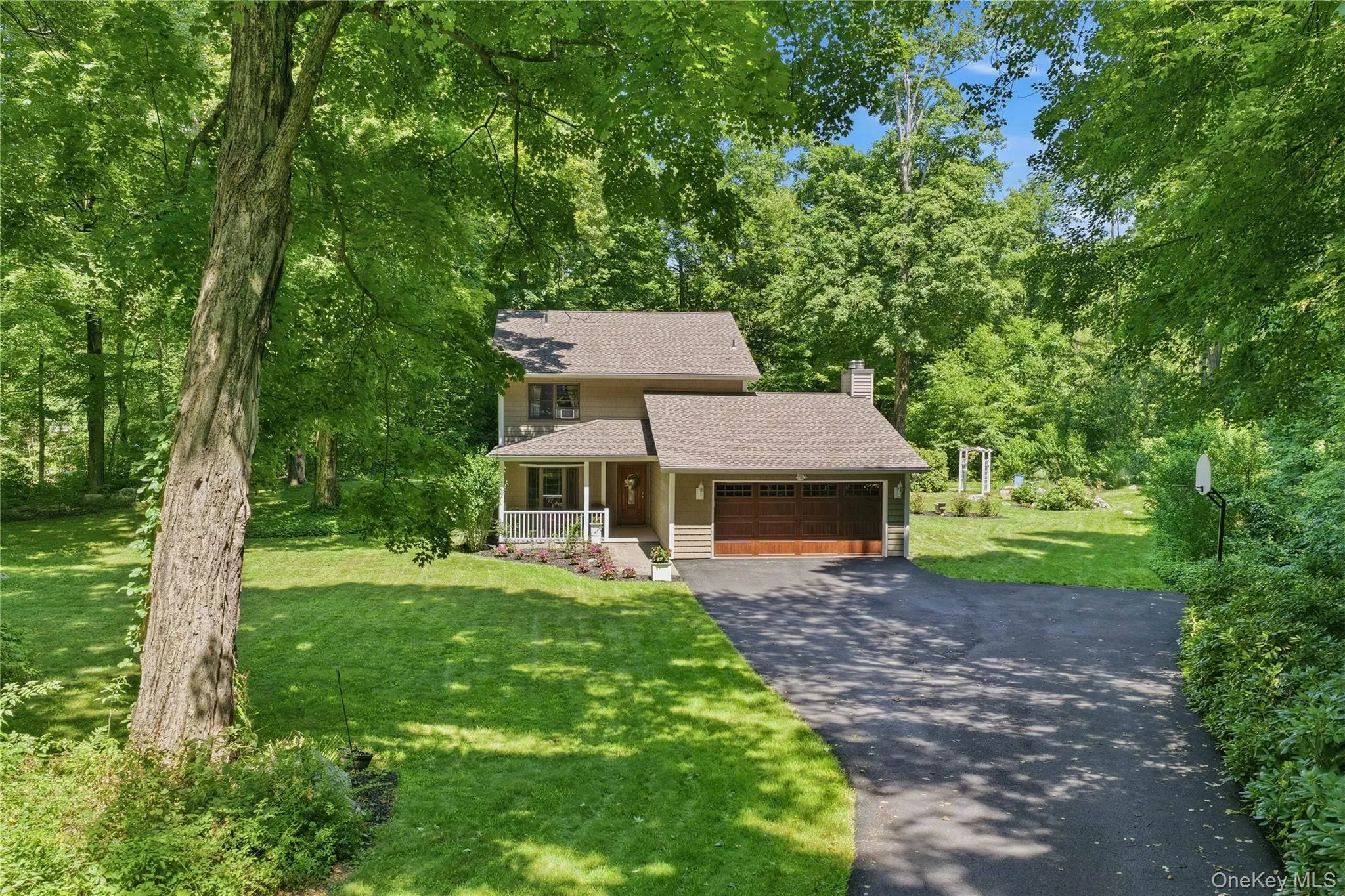 Traditional-style residence featuring a two-car garage with a wood-finish door, a covered front porch with white railings, and a paved driveway Traditional-style residence featuring a two-car garage with a wood-finish door, a covered front porch with white railings, and a paved driveway