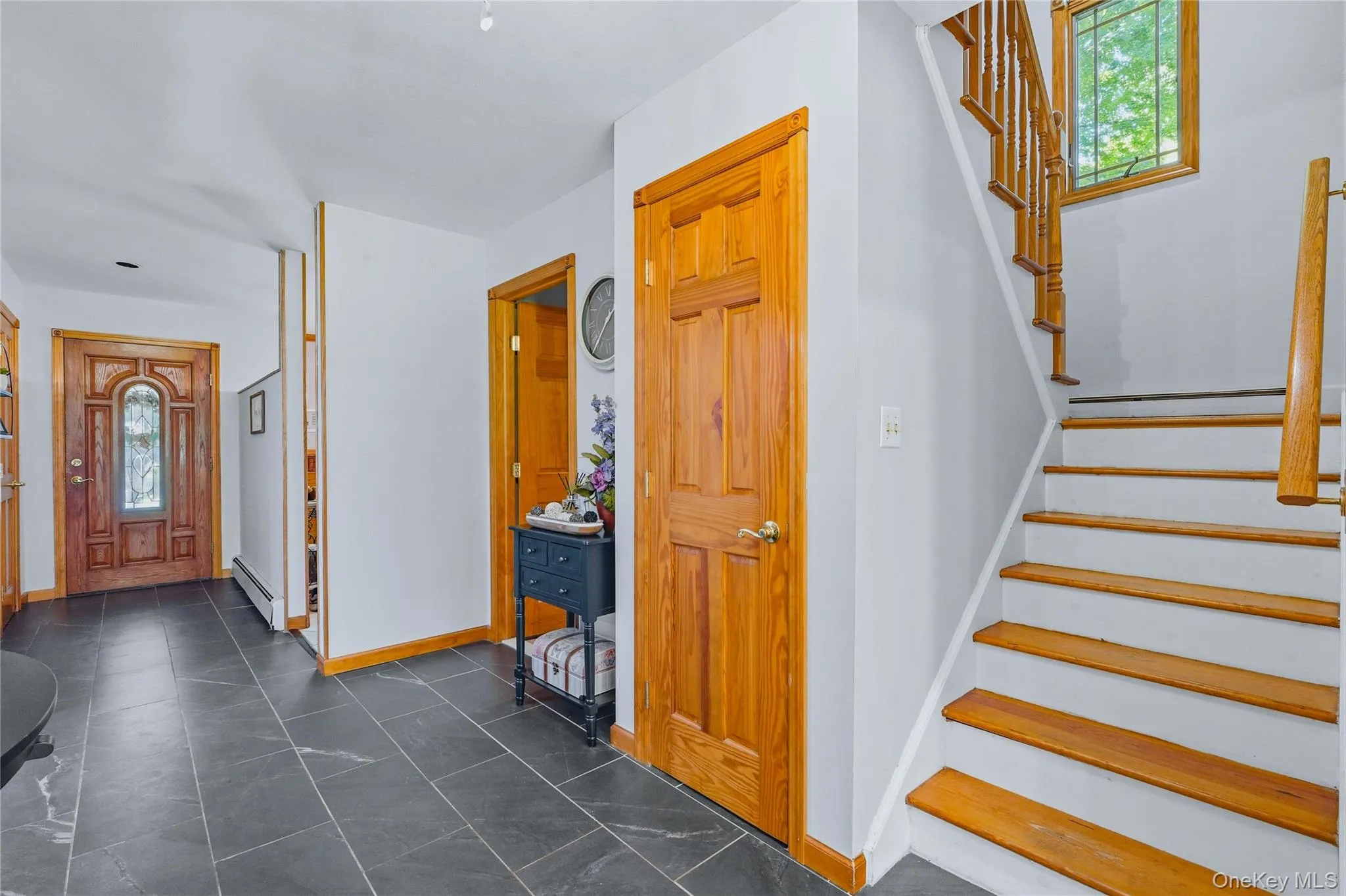 Foyer featuring dark rectangular tile flooring, a decorative wood entry door with leaded glass, a wood staircase with white risers and wood balusters, natural wood interior doors, and white walls with wood trim Foyer featuring dark rectangular tile flooring, a decorative wood entry door with leaded glass, a wood staircase with white risers and wood balusters, natural wood interior doors, and white walls with wood trim