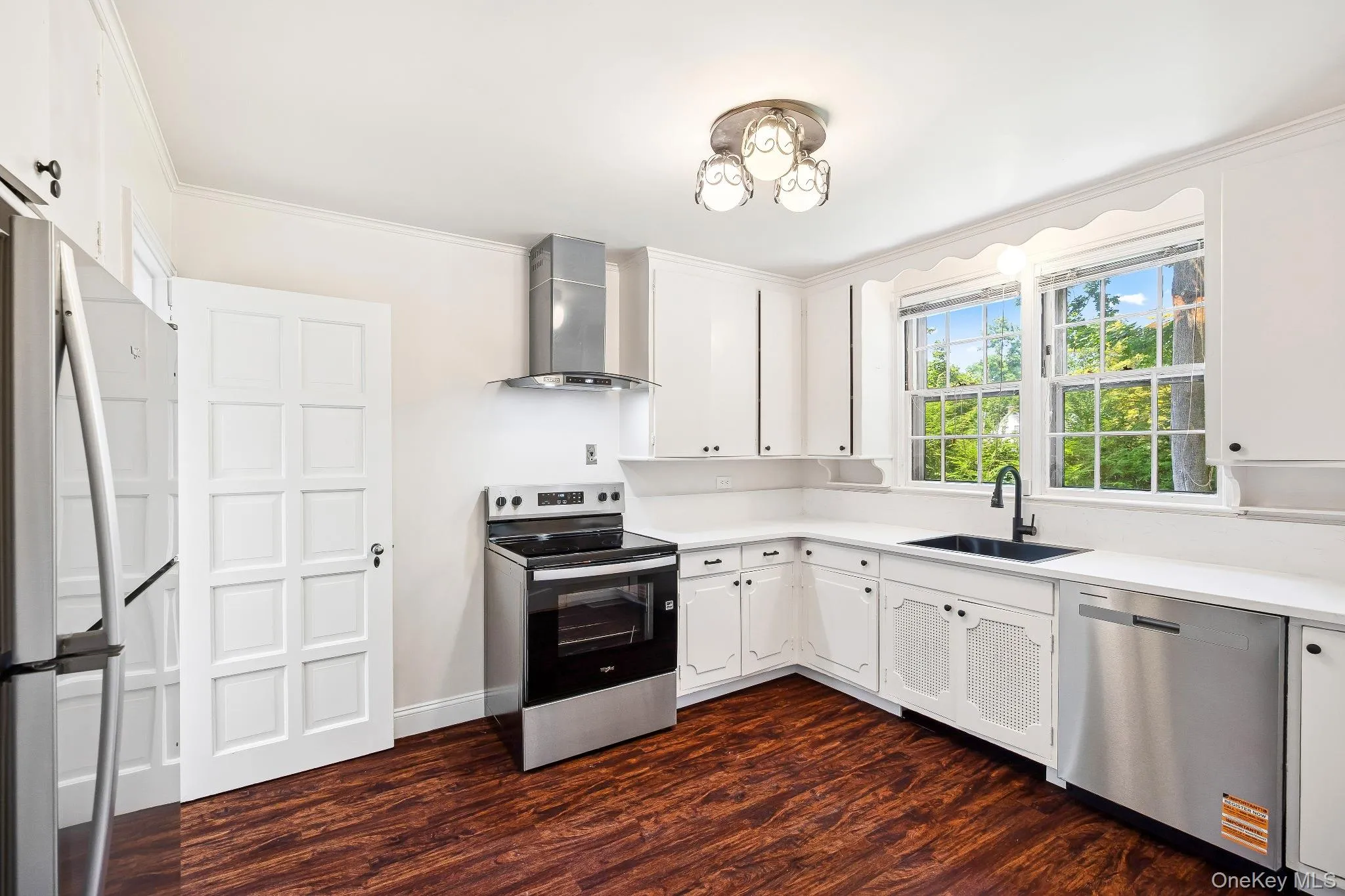 Kitchen featuring appliances with stainless steel finishes, wall chimney range hood, dark wood-style flooring, ornamental molding, and light countertops Kitchen featuring appliances with stainless steel finishes, wall chimney range hood, dark wood-style flooring, ornamental molding, and light countertops