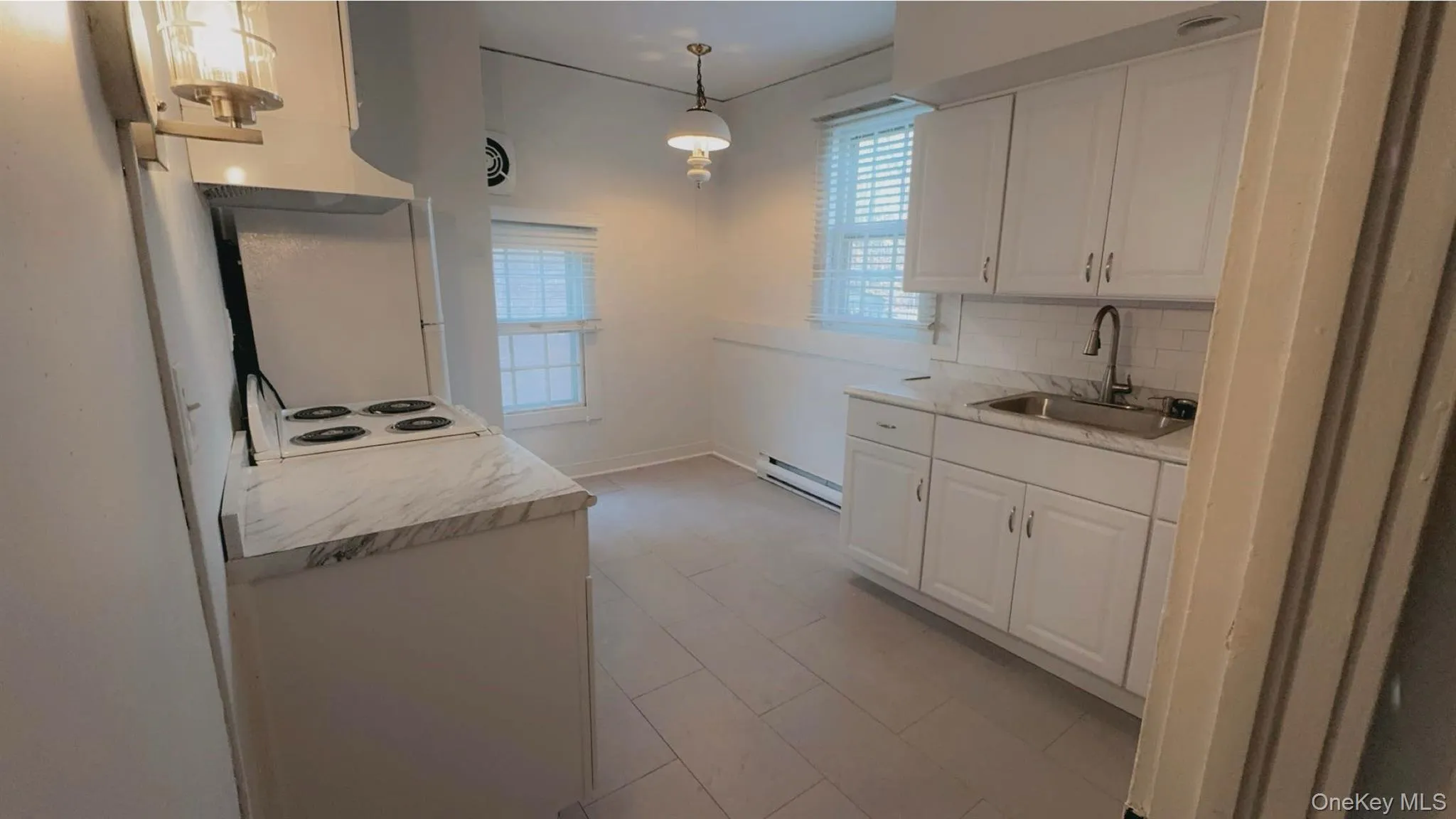 Kitchen with tasteful backsplash, baseboard heating, sink, pendant lighting, and white cabinetry Kitchen with tasteful backsplash, baseboard heating, sink, pendant lighting, and white cabinetry