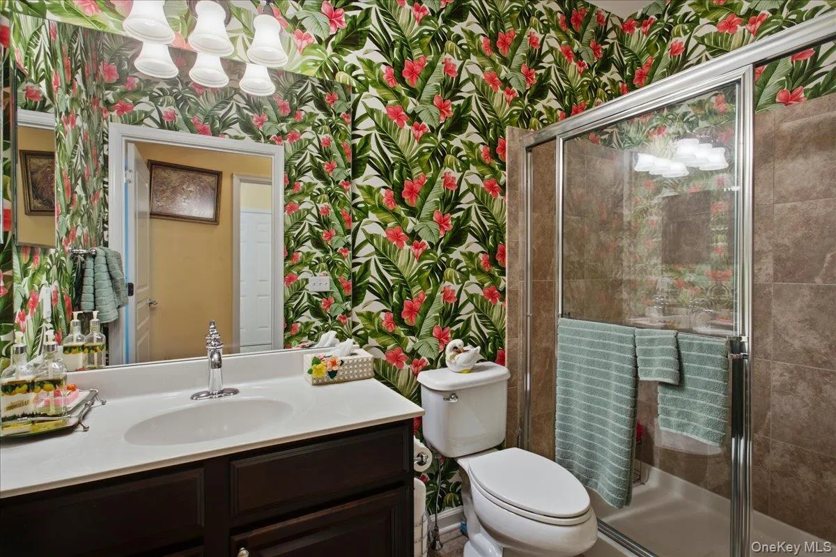 Bathroom featuring vibrant botanical wallpaper, a dark wood-finish vanity with a white countertop and integrated sink, a large mirror, and a shower with glass enclosure and brown tile surround Bathroom featuring vibrant botanical wallpaper, a dark wood-finish vanity with a white countertop and integrated sink, a large mirror, and a shower with glass enclosure and brown tile surround
