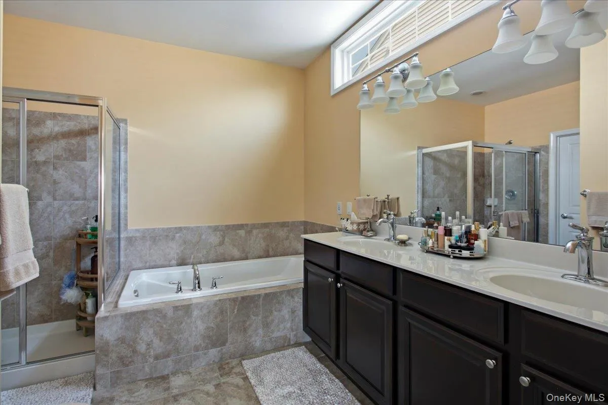 Bathroom featuring a double vanity with dark wood cabinetry and integrated sinks, a built-in soaking tub with stone-finish tile surround, a glass-enclosed shower, and a high transom window Bathroom featuring a double vanity with dark wood cabinetry and integrated sinks, a built-in soaking tub with stone-finish tile surround, a glass-enclosed shower, and a high transom window