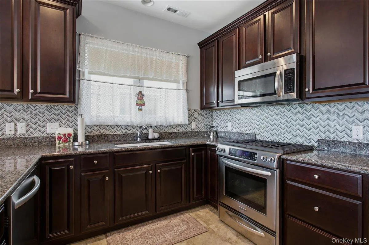 Corner kitchen with dark wood cabinetry, stainless steel appliances, granite countertops, and a chevron-patterned tile backsplash Corner kitchen with dark wood cabinetry, stainless steel appliances, granite countertops, and a chevron-patterned tile backsplash