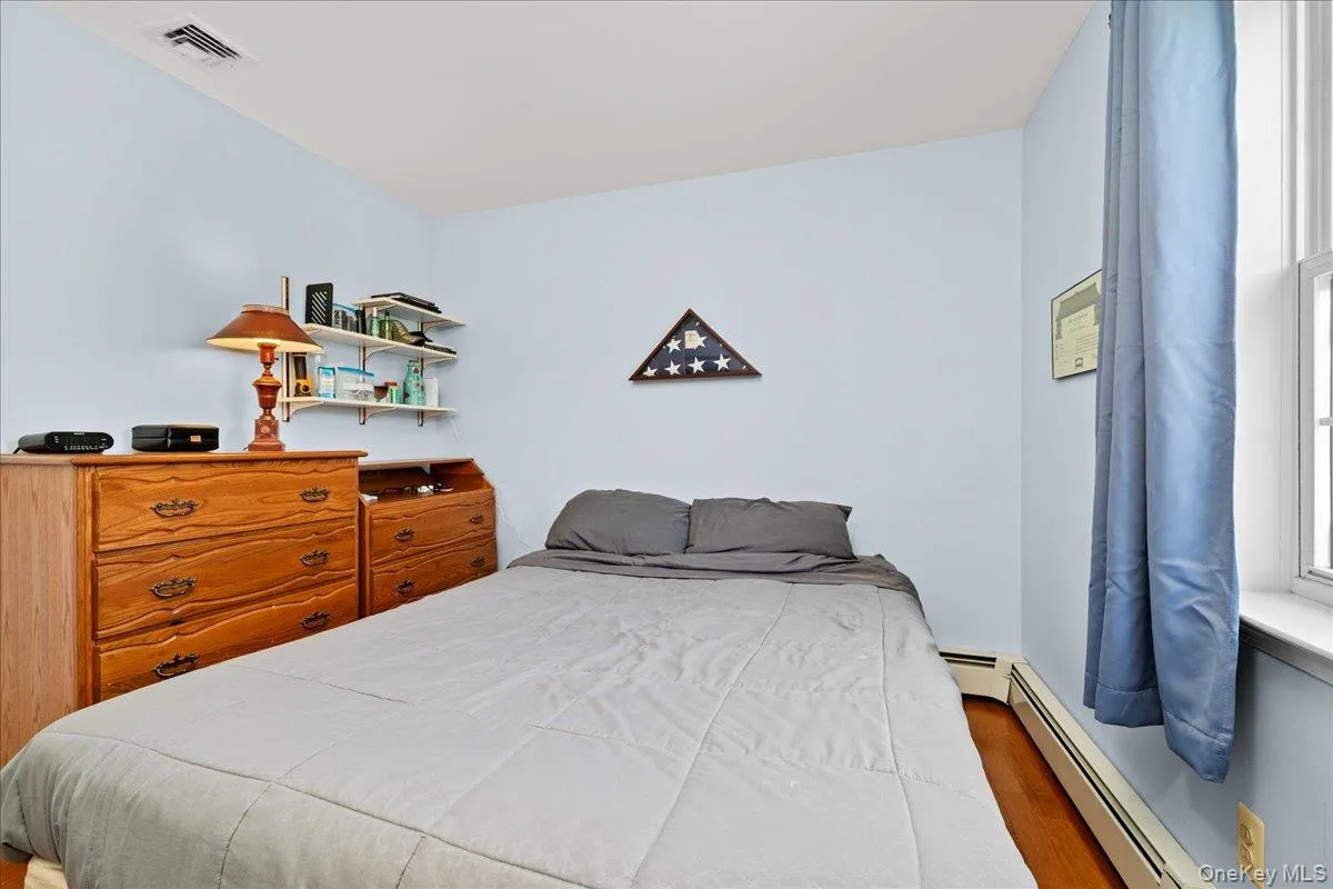 Bedroom 2 featuring hardwood flooring, light blue wall paint, a window with natural light, a baseboard heating unit, and a ceiling vent Bedroom 2 featuring hardwood flooring, light blue wall paint, a window with natural light, a baseboard heating unit, and a ceiling vent