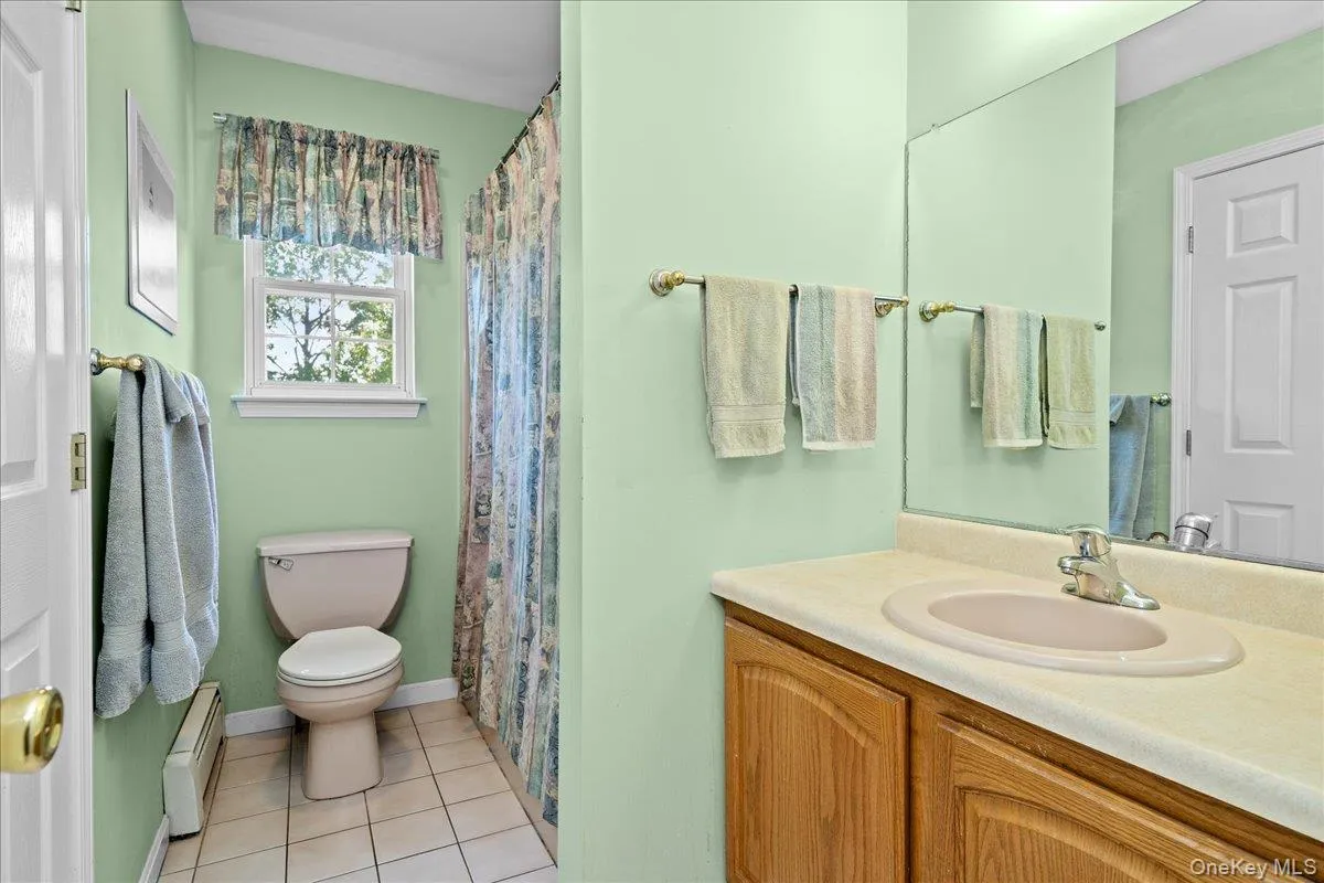 Full bathroom featuring a single-sink vanity with wood-finish cabinetry, large wall-mounted mirror, tiled flooring, a window, and a built-in toilet Full bathroom featuring a single-sink vanity with wood-finish cabinetry, large wall-mounted mirror, tiled flooring, a window, and a built-in toilet