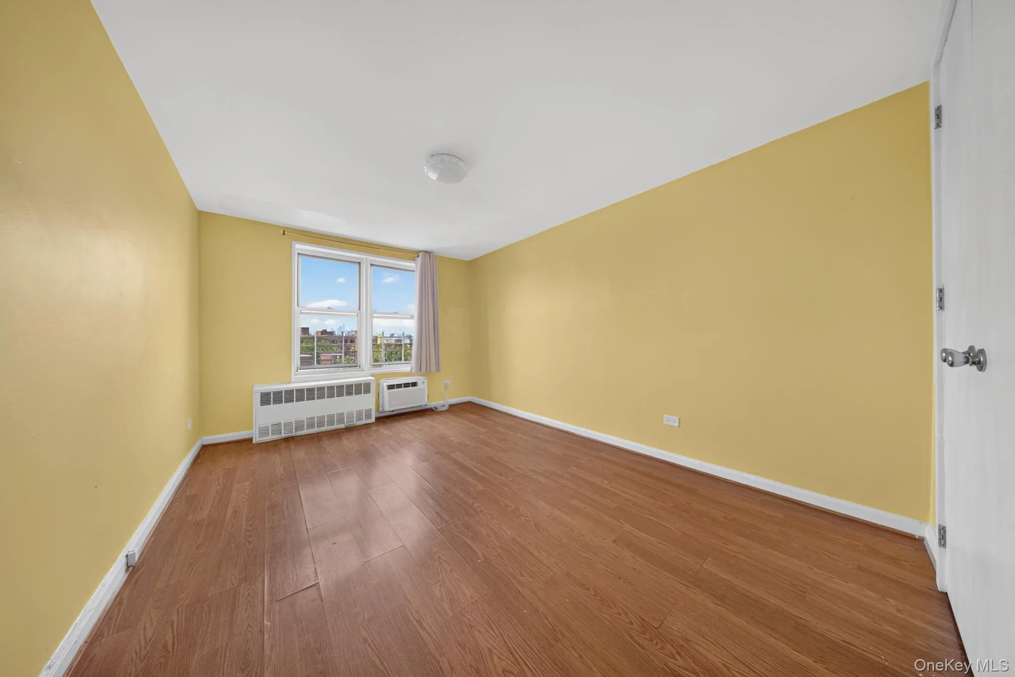 Spacious room featuring wood-finish flooring, a large window, and light yellow walls Spacious room featuring wood-finish flooring, a large window, and light yellow walls